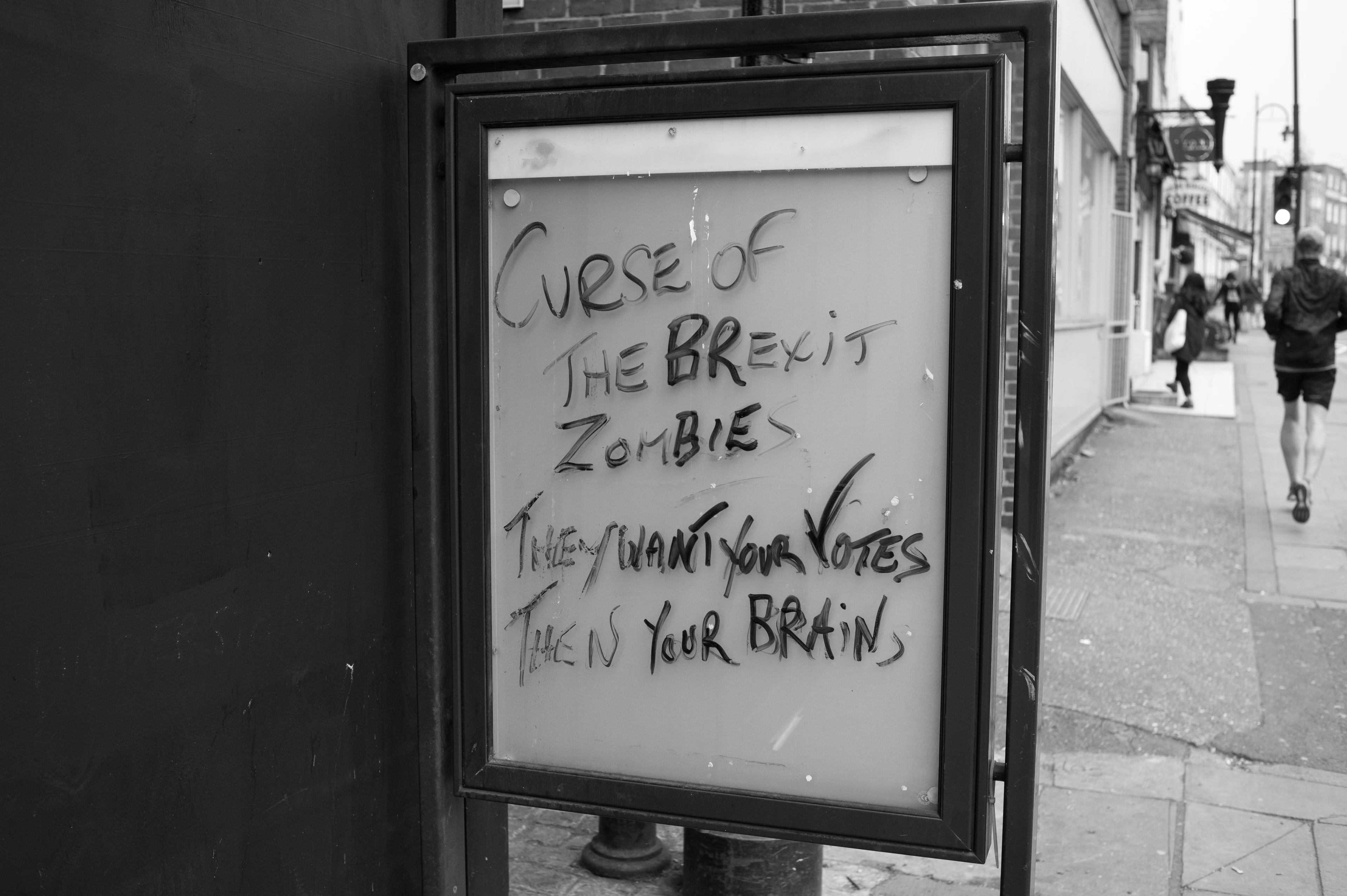 Black-and-white image: A protest message written in felt-tip pen on the glass of a poster display case at a bus stop with the words ‘Curse of the Brexit Zombie – They want your votes, then your brains’.