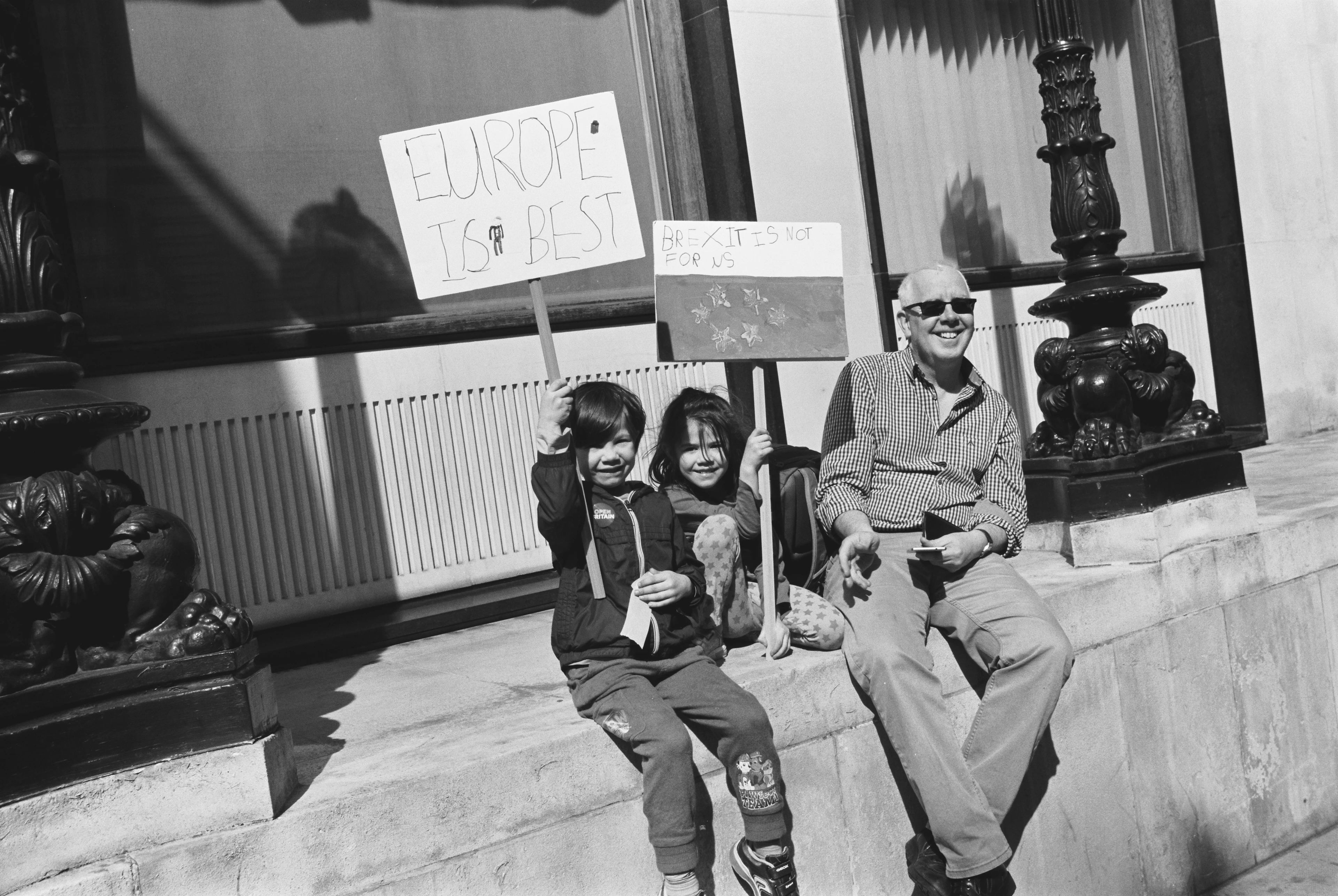 Black-and-white image: Two children and an elderly man sit relaxed in the sun at the side of the road. One child holds up a protest sign saying ‘Europe is best’, and the other holds one saying ‘Brexit is not for us’. The man smiles next to them.