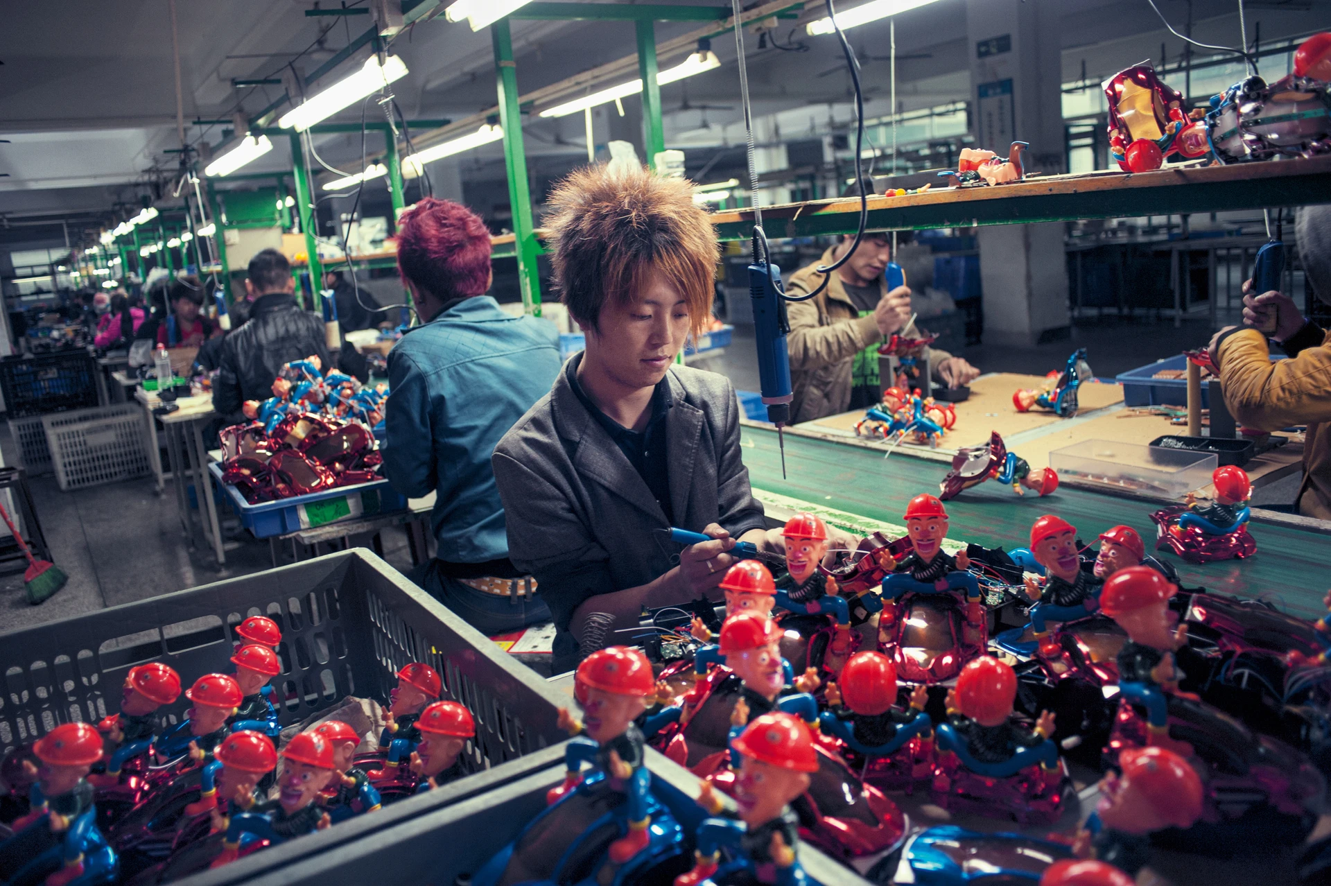 Arbeiterinnen und Arbeiter in einer Fabrik montieren bunte Spielzeugfiguren an langen Produktionstischen unter grellem Neonlicht.