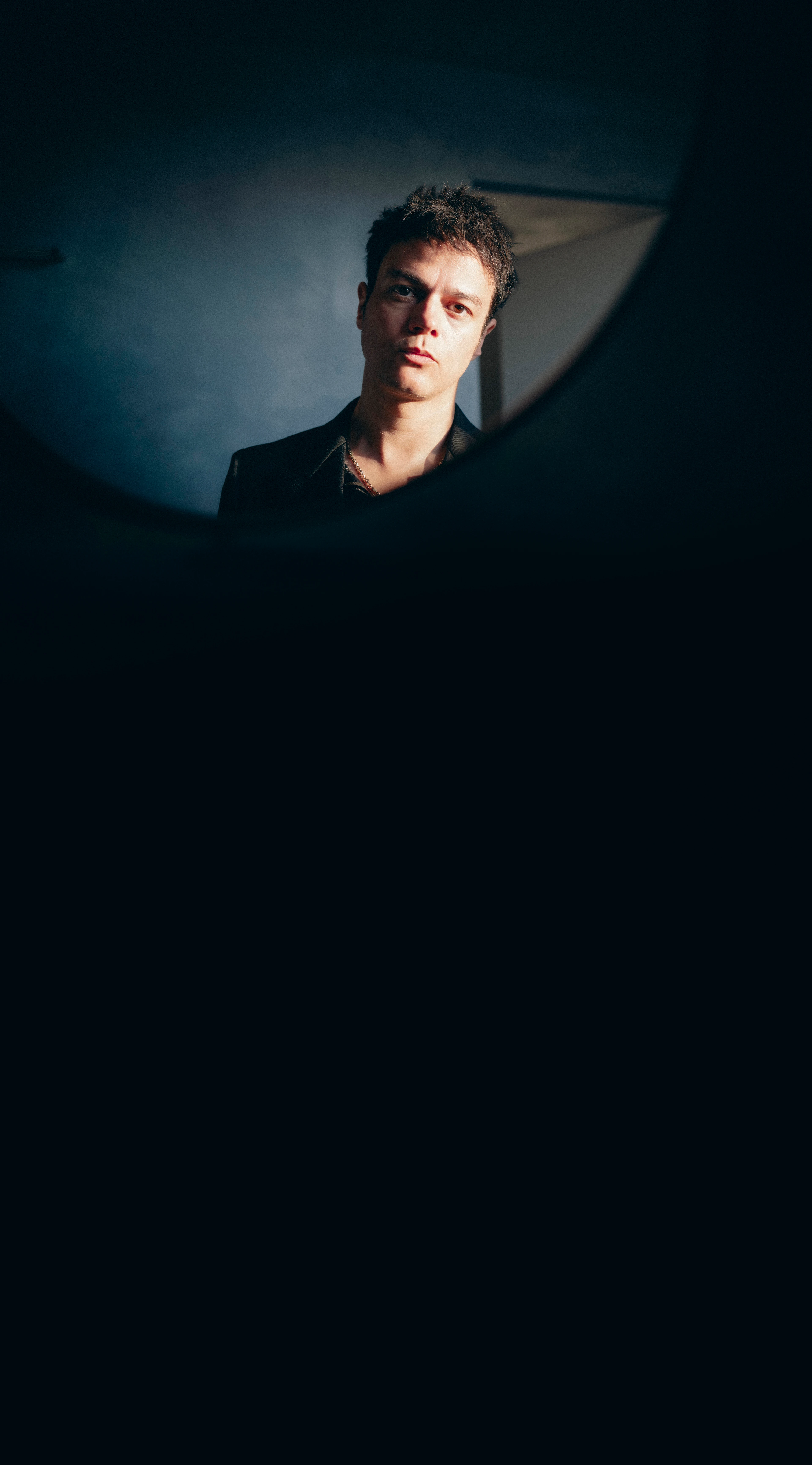 Jamie Cullum, con expresión seria, aparece reflejado en un espejo; el fondo parece oscuro y suavemente iluminado, lo que crea un tono reflexivo e íntimo.