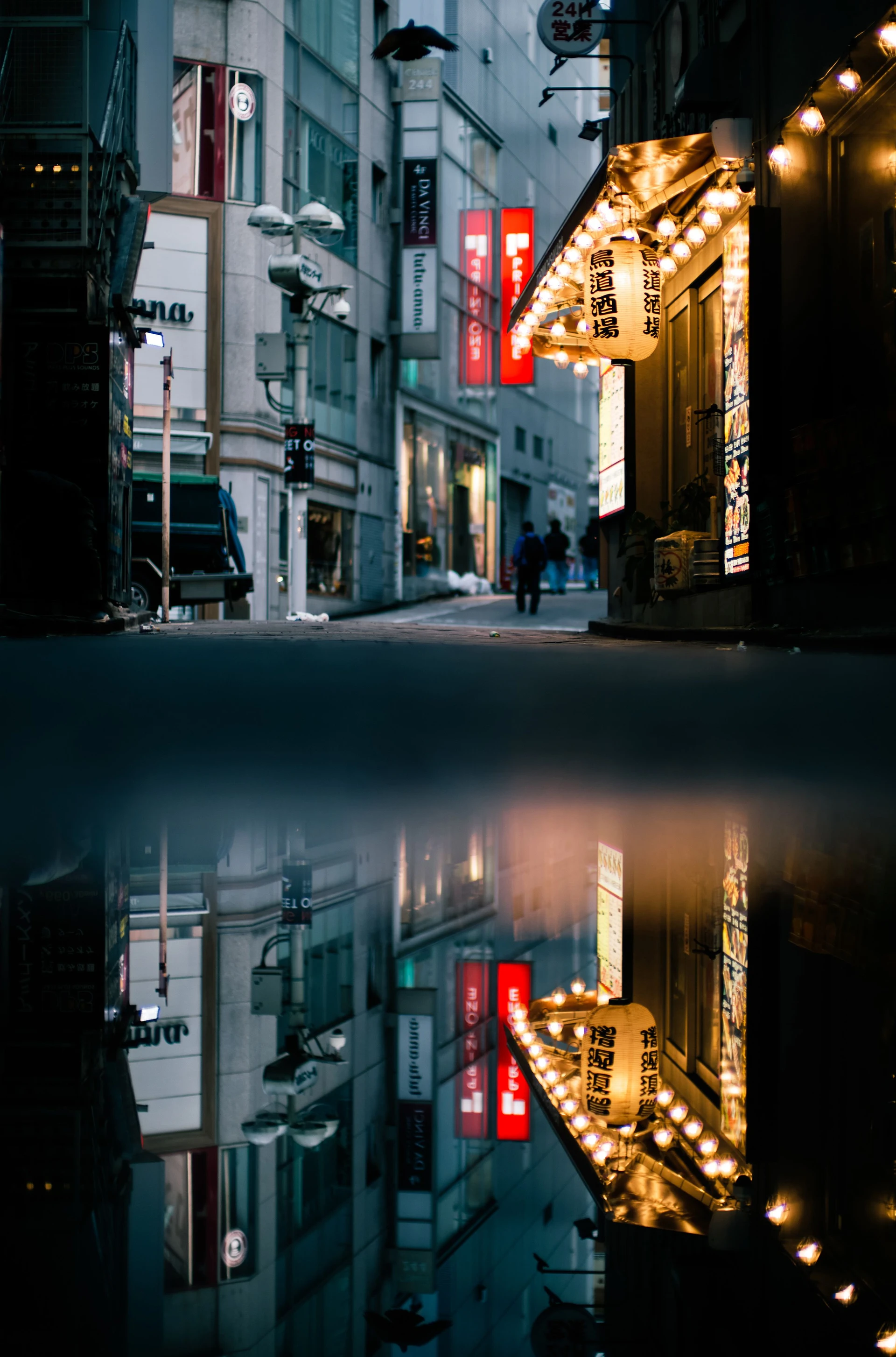 A Tokyo street scene at blue hour: The reflection of a dark alleyway is doubled in a puddle, directing our gaze towards facades with a conspicuously illuminated restaurant and a red sign.