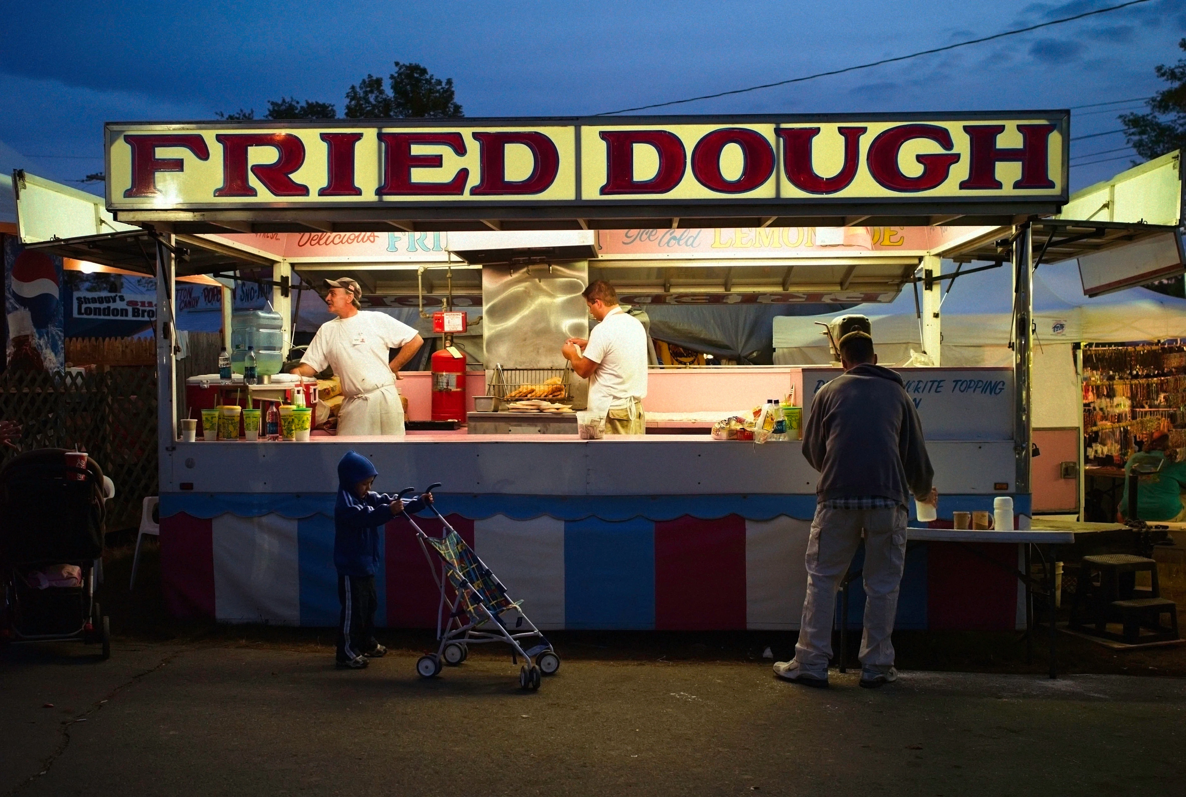 Atmosferyczne spojrzenie na oświetloną budkę z przekąskami w parku rozrywki o zmierzchu, przed którą stoi dwóch pracowników w białych strojach i dziecko z ojcem.