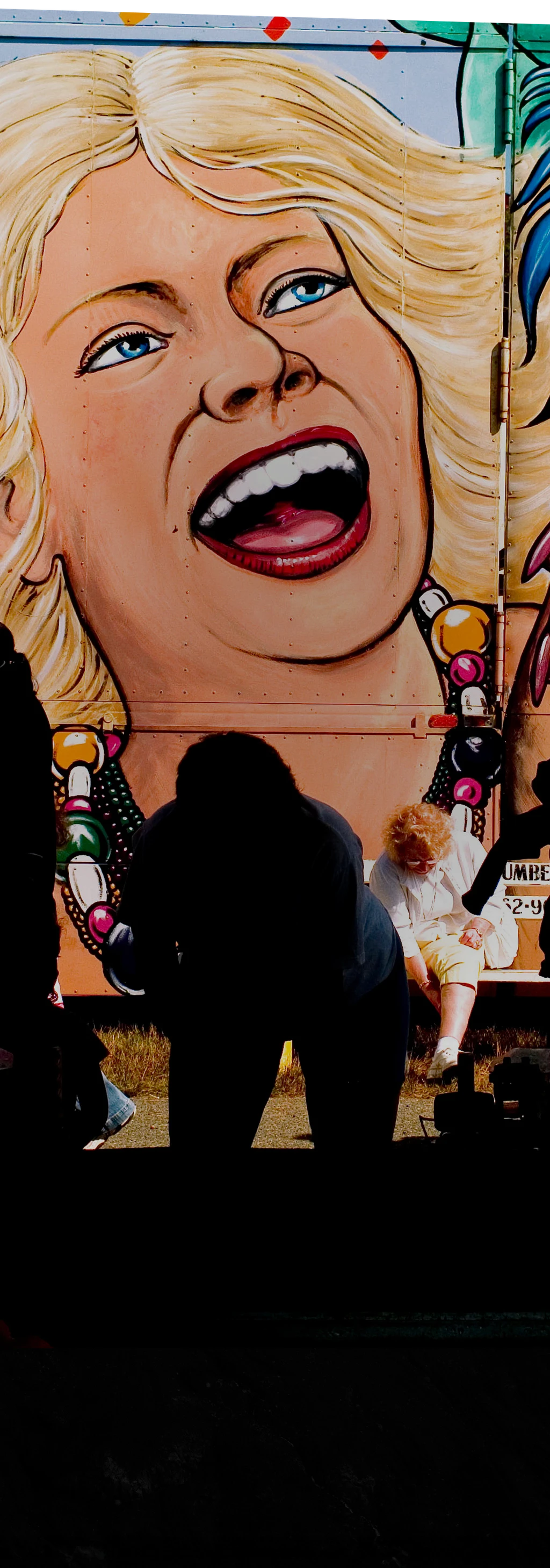 Kolorowy fragment muralu przedstawiający blondynkę trzymającą maskę karnawałową, przed którą stoją dwie przechodnie.