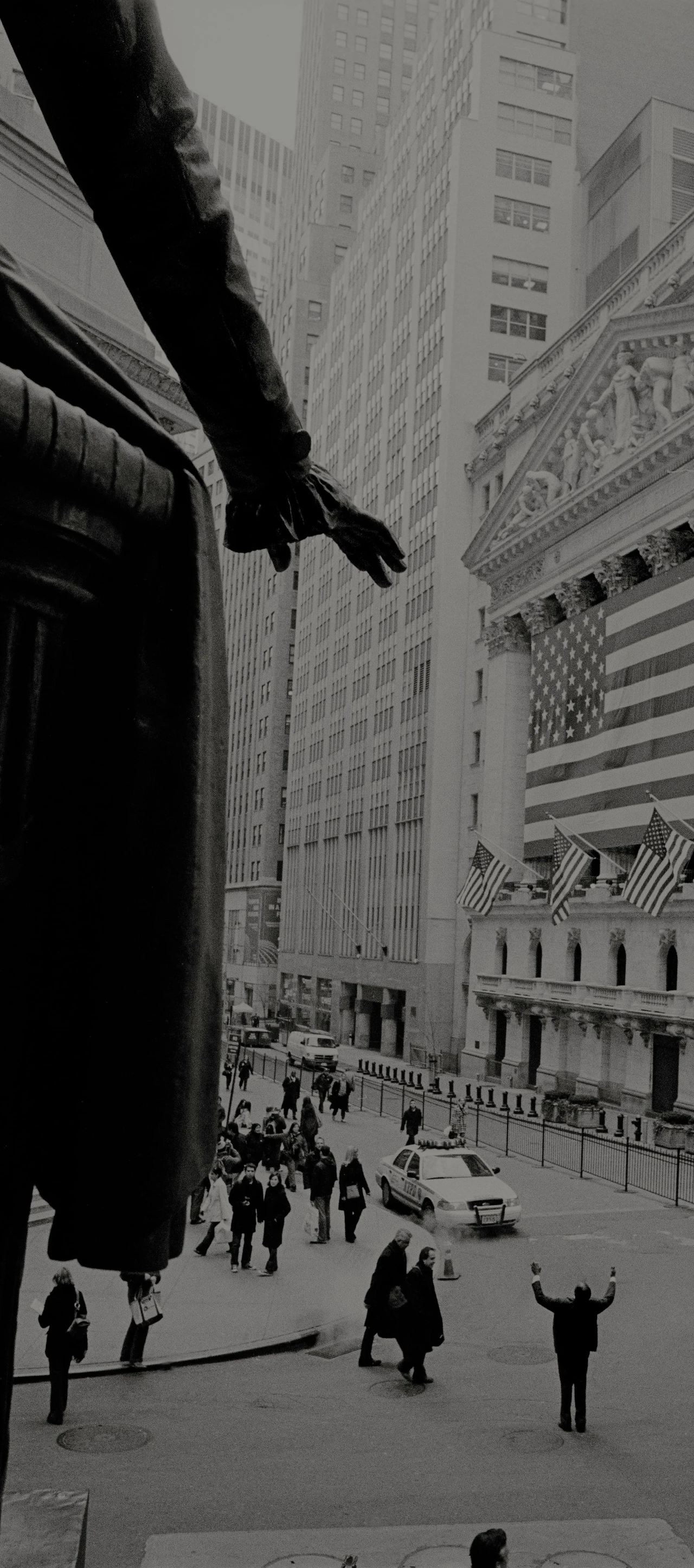 Imponujące czarno-białe zdjęcie przedstawiające pokrytą amerykańskimi flagami fasadę nowojorskiej giełdy papierów wartościowych, przed którą mężczyzna modlący się wśród przechodniów podnosi rękę.