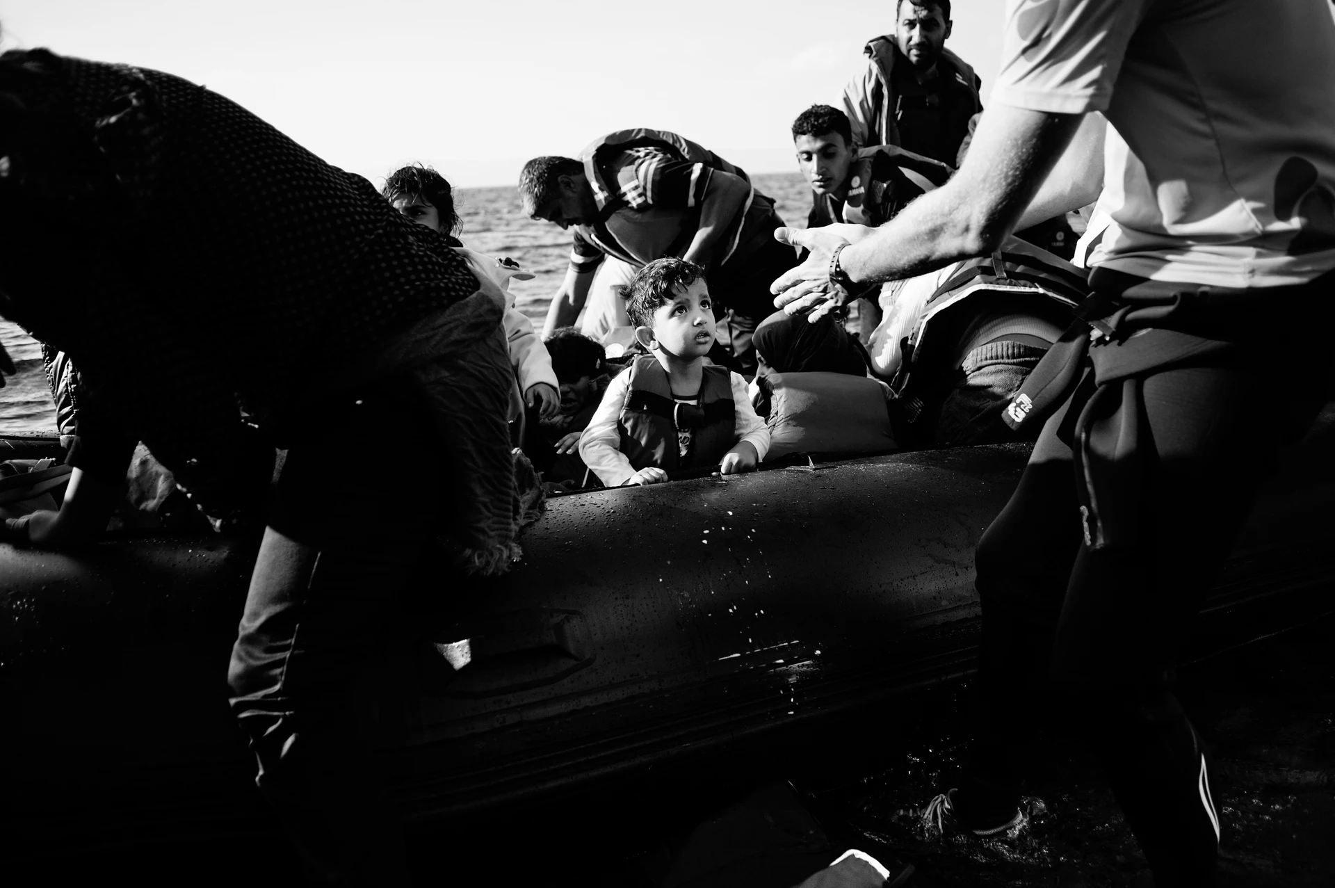 Several people in shallow water work together to pull an overcrowded rubber dinghy to the shore. In the centre of the picture, a small boy looks wide-eyed in the direction of a helper.