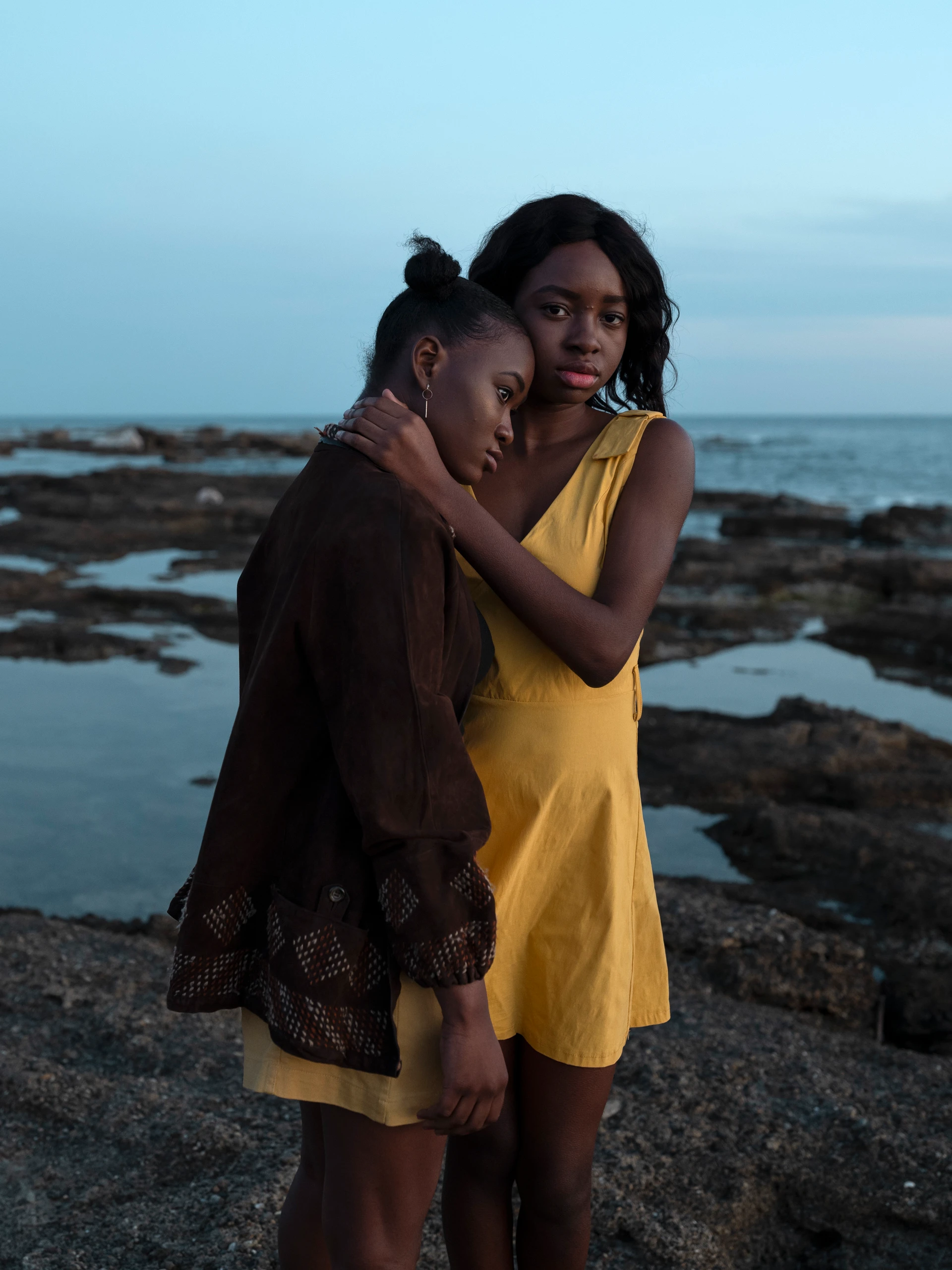 Two young Black women stand close together on a rocky stretch of coastline. One of them is wearing a yellow dress and is embracing the other woman wearing a brown coat over a yellow dress. She is looking forwards while the other rests her head on the first woman’s shoulder.