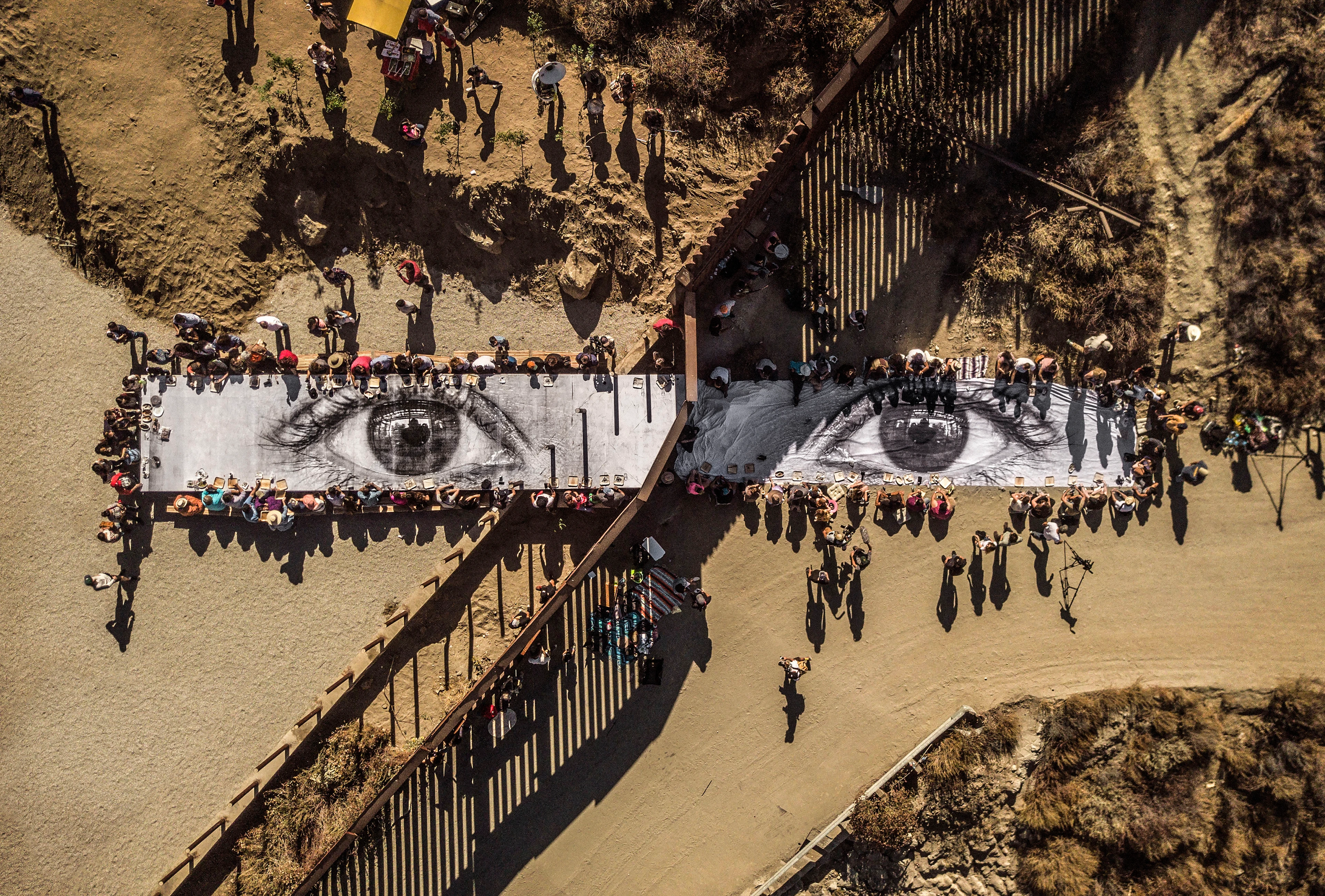 Bird’s eye view: A large group of people sit and eat together at a table and on a blanket. Through a border fence, they form a common image of two eyes looking up at the sky.