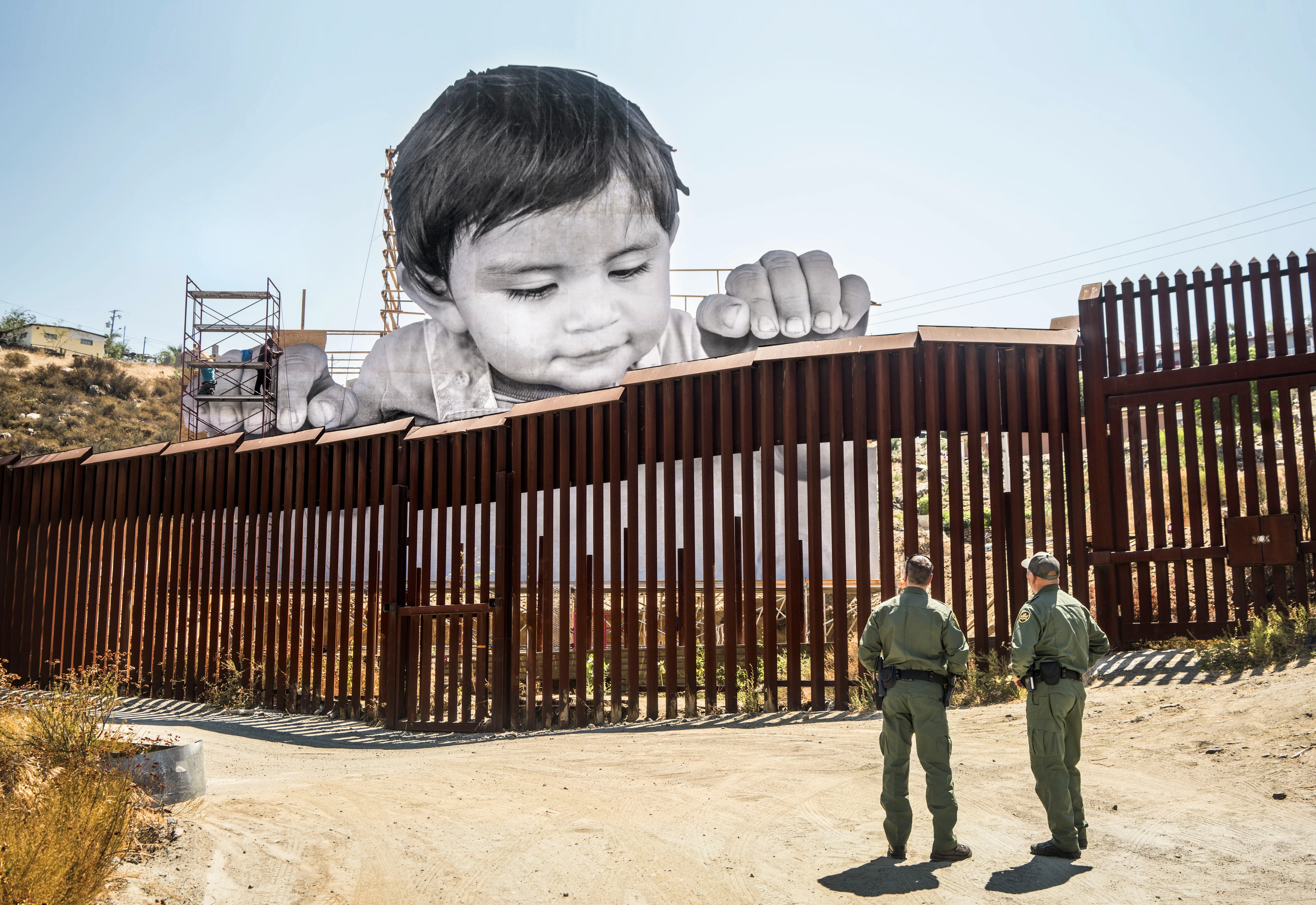 A large-format image of a child looking over a border fence and placing their hands on it stretches across the top of the fence, while two uniformed officers stand in front of it.