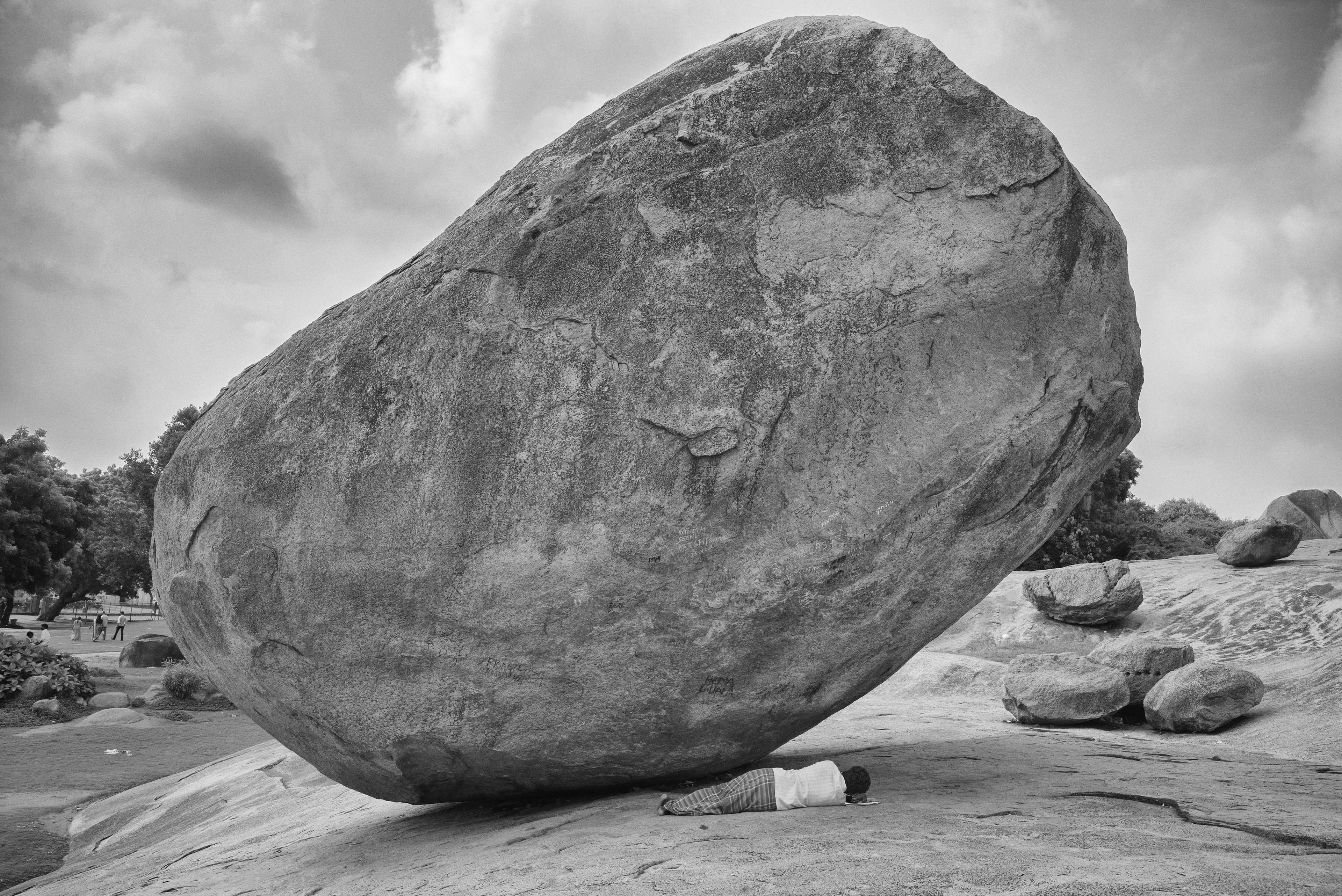 Black-and-white image: A large, rounded boulder leans at an angle on a rocky surface in the open air. In front of it, in its shadow, a relatively small person lies sleeping.