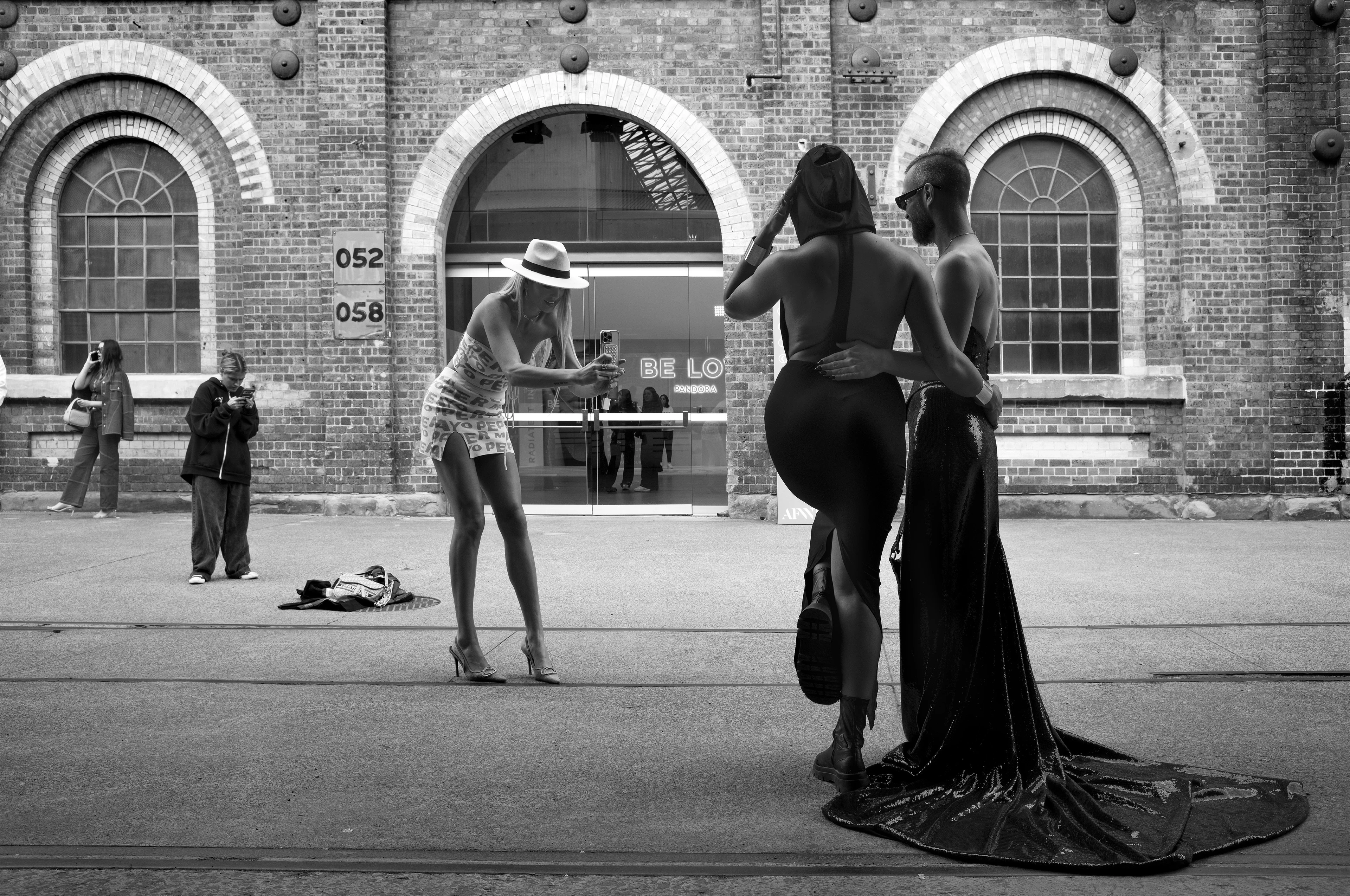 Black-and-white image: A man and a woman, both wearing dresses, pose together in a square in front of a brick building. A woman wearing a sun hat, a tight white dress and high heels is photographing them. In the background, two other women are busy with their smartphones.