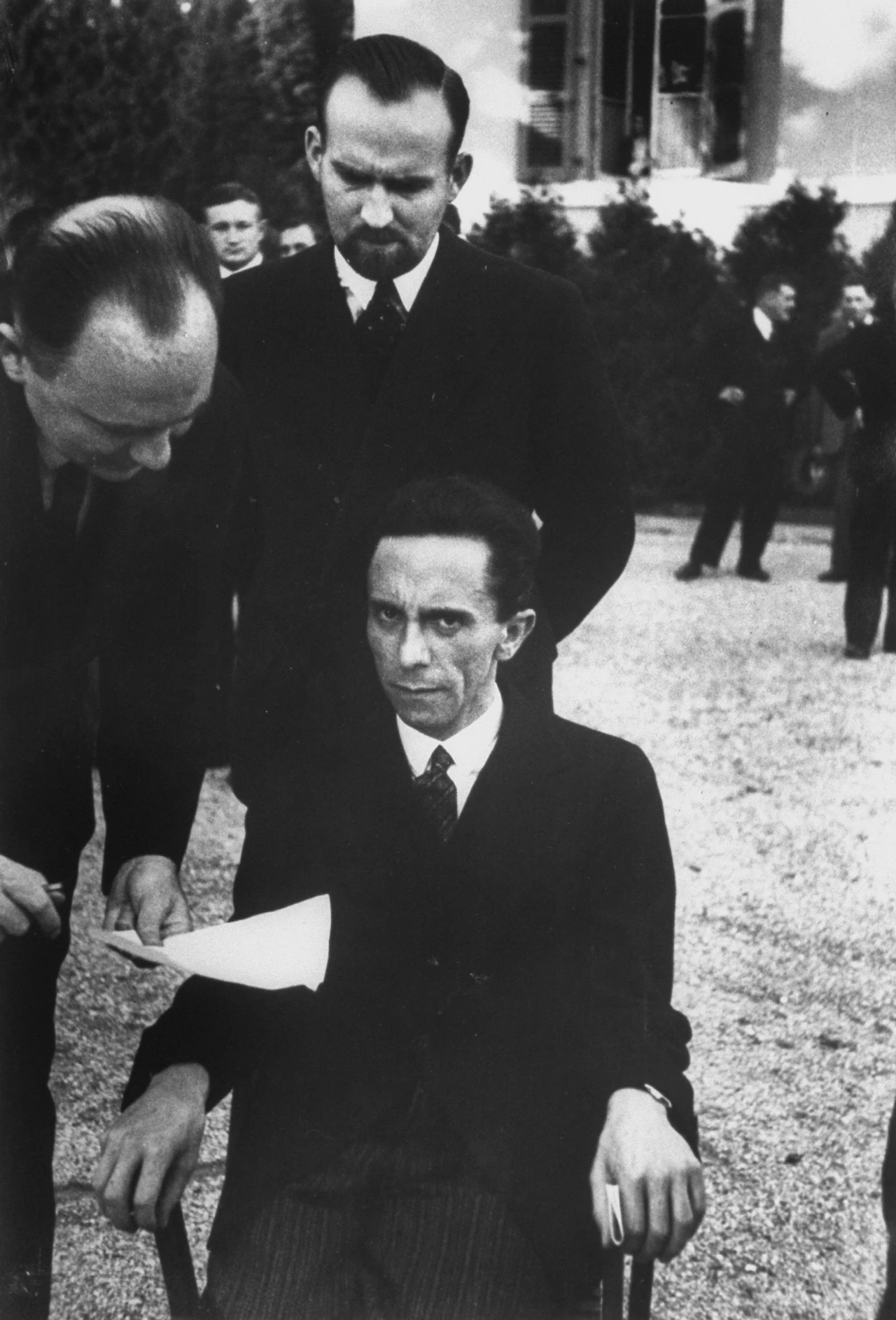 黑白照片，一名男子坐在椅子上，双手紧抓椅背，神情阴沉。背景中站着两名男子。