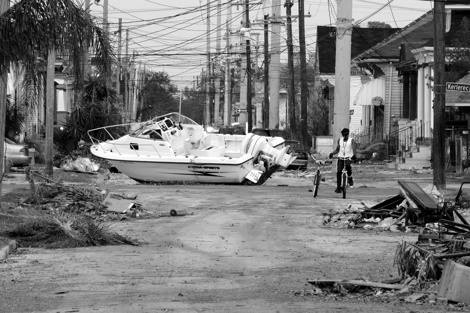 Schwarz-Weiß-Bild mit Blick in die Flucht einer verwüsteten Straße in New Orleans auf der ein Sportboot liegt an dem ein Mann mit zwei Fahrrädern vorbeifährt.
