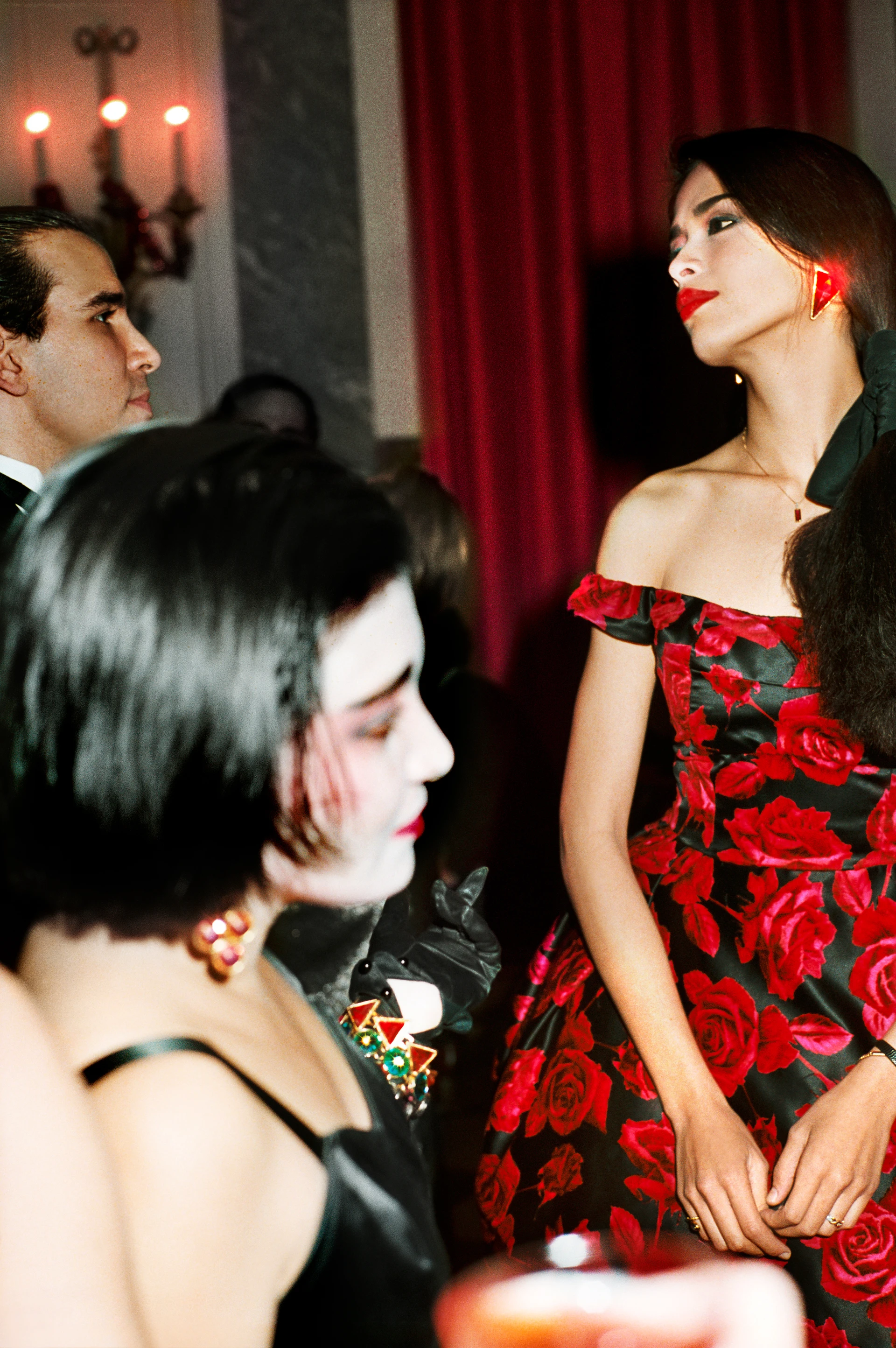 Escena durante el descanso de un baile, una mujer destaca con su vestido de rosas rojas y un pendiente rojo brillante en la oreja derecha; a la izquierda, otras dos personas vestidas elegantes.