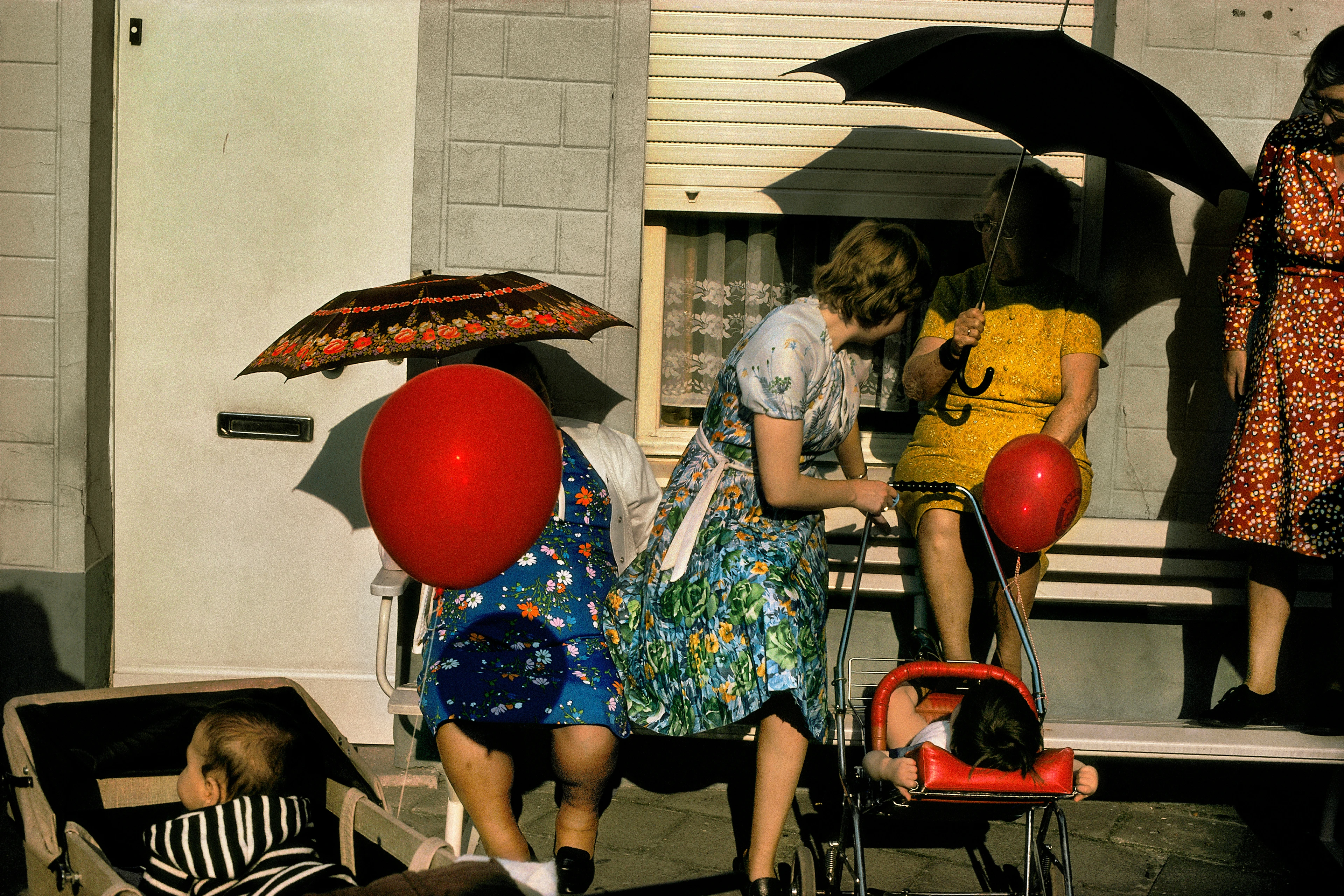 Escena extravagante: madres con vestidos de flores y paraguas y dos bebés en carritos con globos rojos se mueven frente a un edificio de viviendas gris.