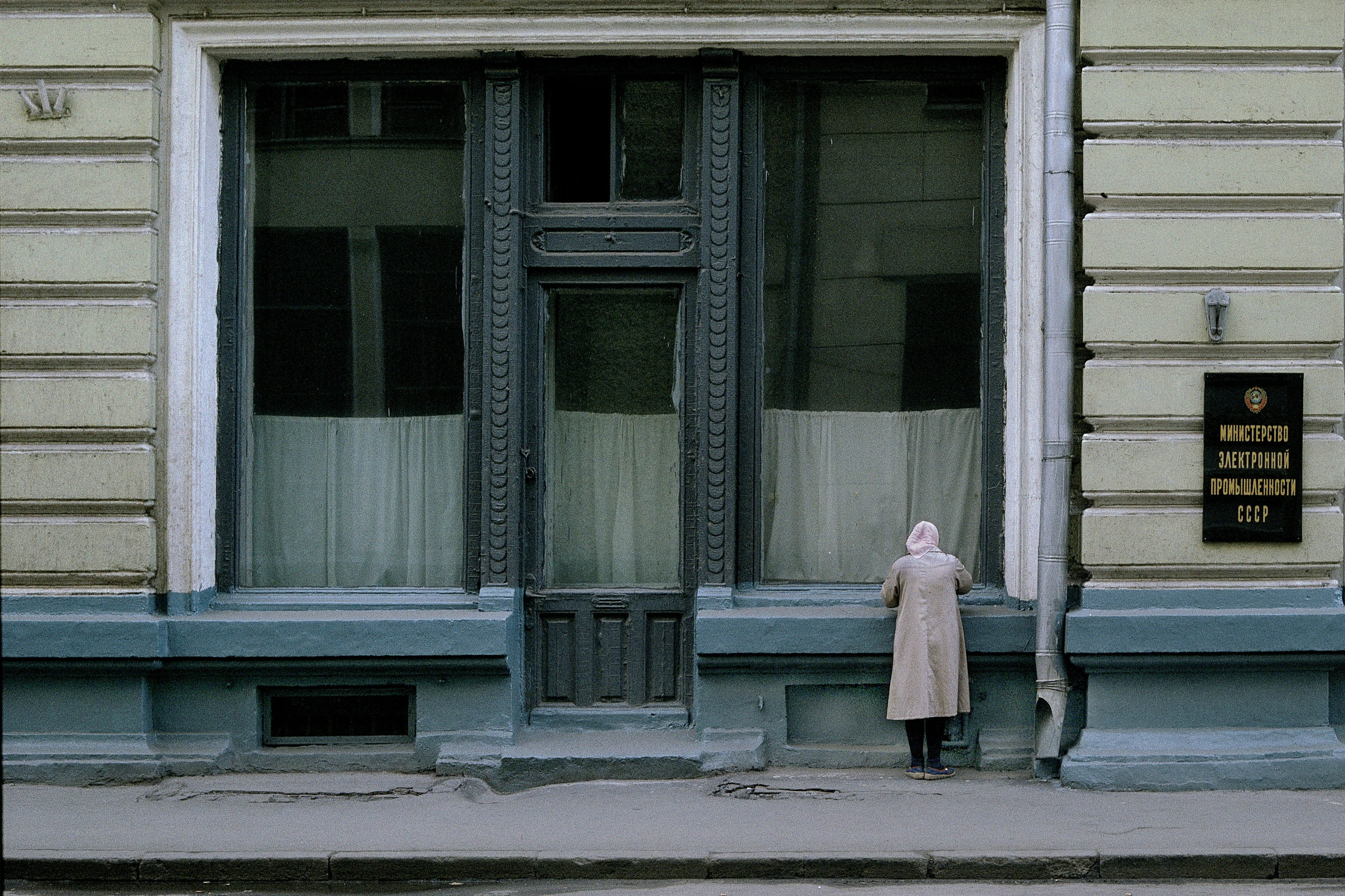 Una mujer con parka y pañuelo en la cabeza se encuentra delante de una gran ventana con cortinas en un escenario soviético gris.