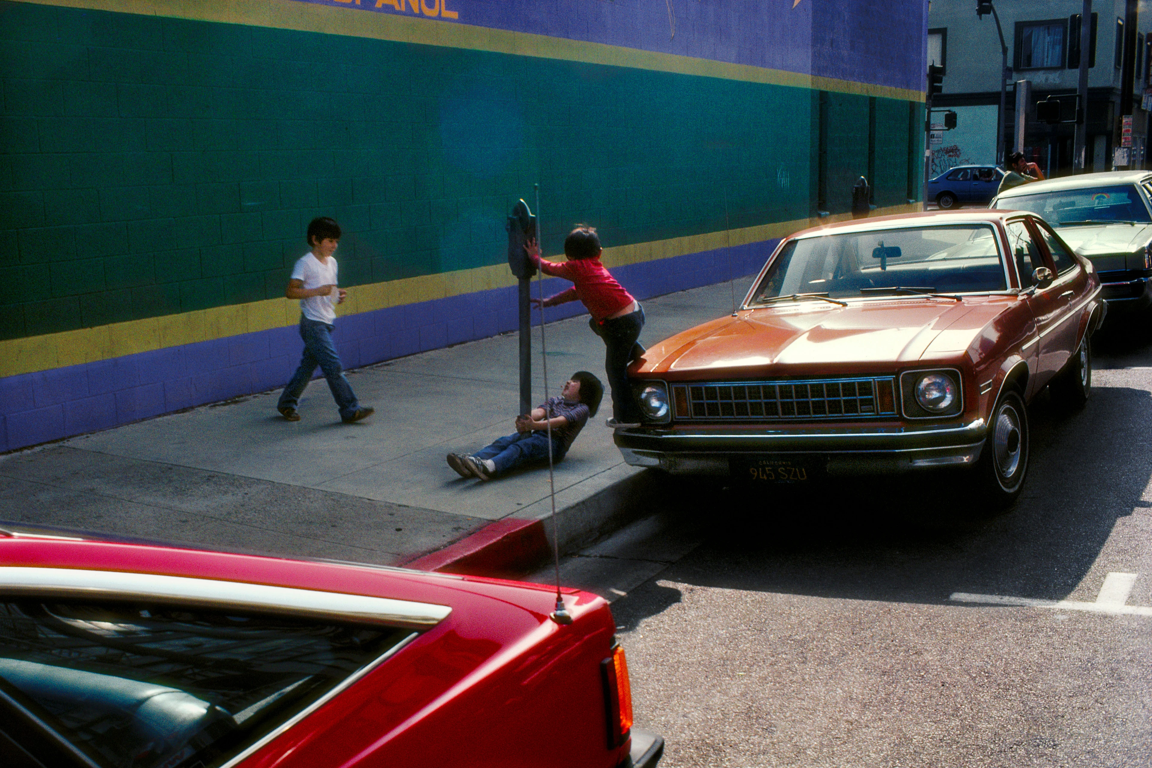 Fotografía con coches y colores típicos de la década de 1980, que muestra tres chicos jugando en la acera con un parquímetro. 