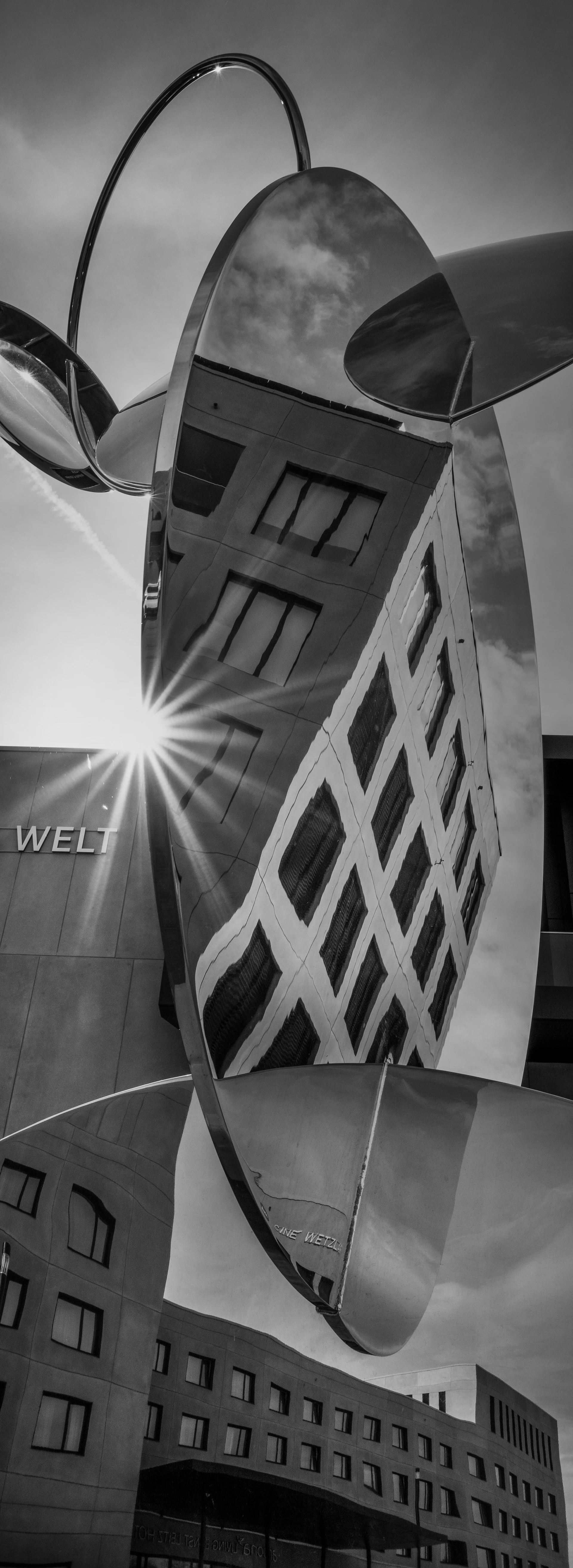 黑白照片:一个严重变形的镜面物体反射着周围的建筑和低垂的太阳,建筑立面上可以看到“Leica Welt”的字样。