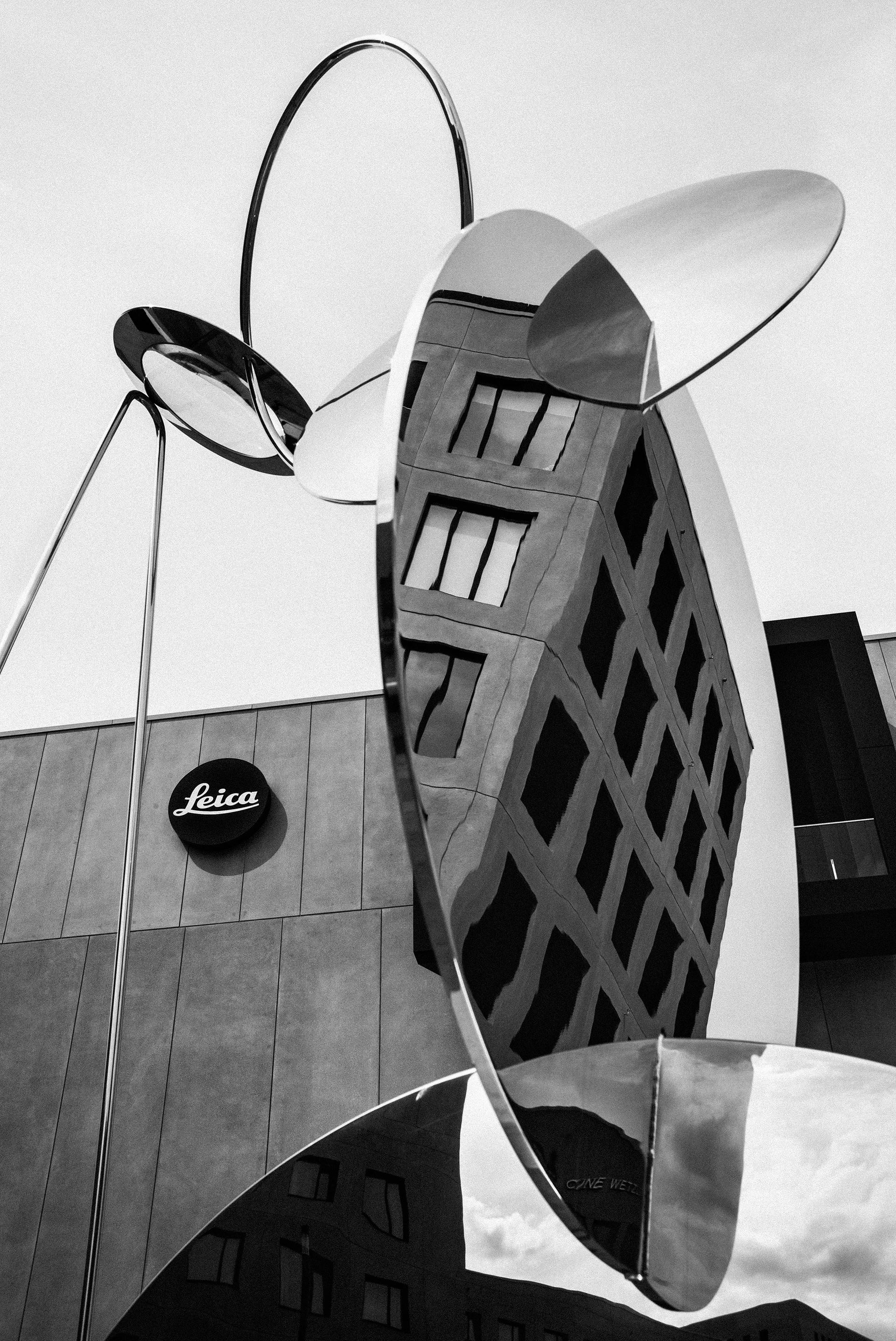 Black-and-white image: A large, reflective metal sculpture distorts the façade of a building, with the Leica logo visible in the background. Curved elements of the sculpture reach up into the sky.