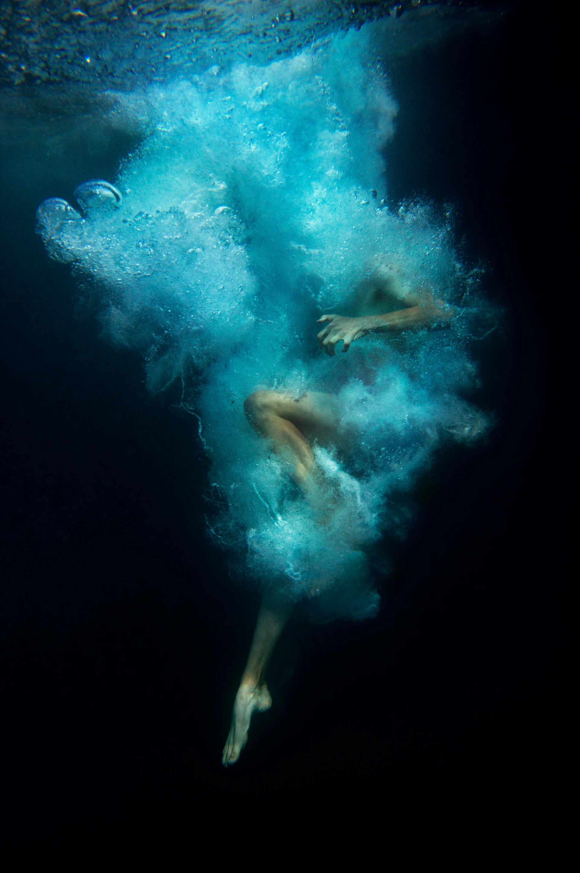 Eine Person taucht ins dunkle Wasser ein, ihr Körper ist von einer großen Wolke aus Luftblasen und Verwirbelungen umhüllt, sodass nur Beine und ein Arm schemenhaft sichtbar sind.