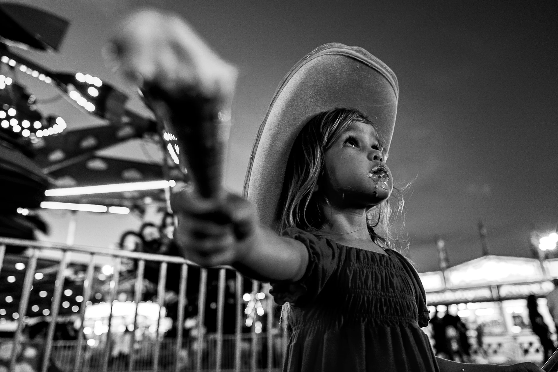 Schwarz-Weiß-Aufnahme: Ein Kind mit großem Cowboyhut hält ein Eis in den Vordergrund und schaut nach oben, während im Hintergrund ein beleuchteter Jahrmarkt unscharf leuchtet.
