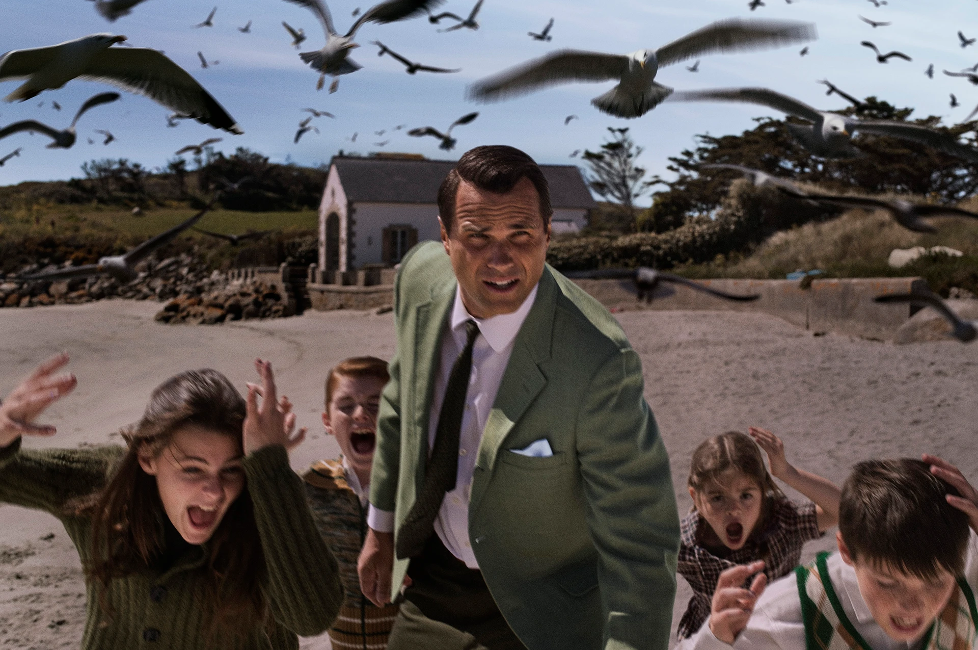 Nachstellung einer berühmten Filmszene: Eine Gruppe von Kindern und ein Mann im Anzug ducken sich erschrocken am Strand, während zahlreiche Vögel tief über sie hinwegfliegen.
