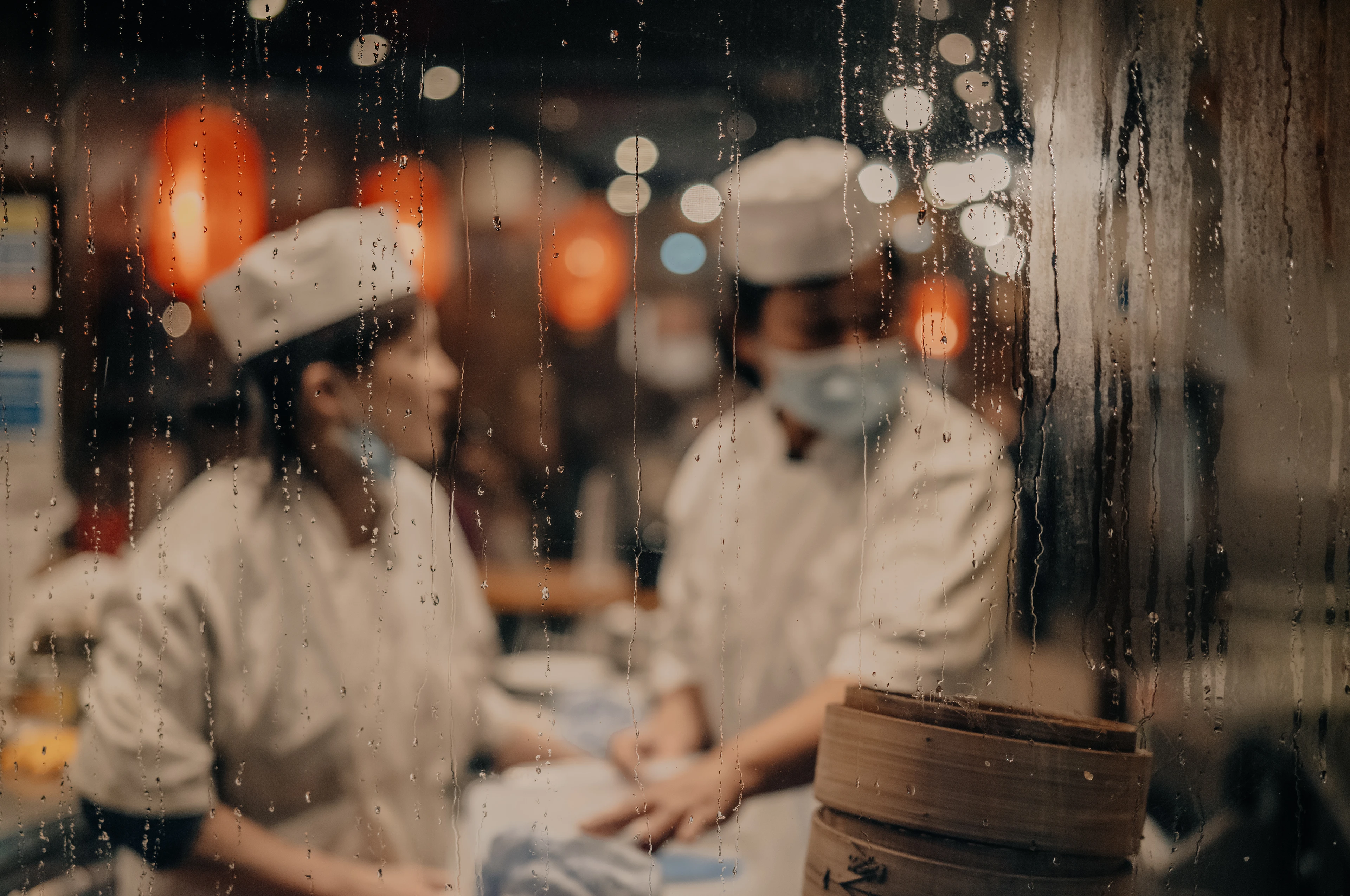 Eine Köchin und ein Koch mit Mundschutz arbeiten hinter einer beschlagenen Glaswand in warmem, unscharfem Restaurantlicht.