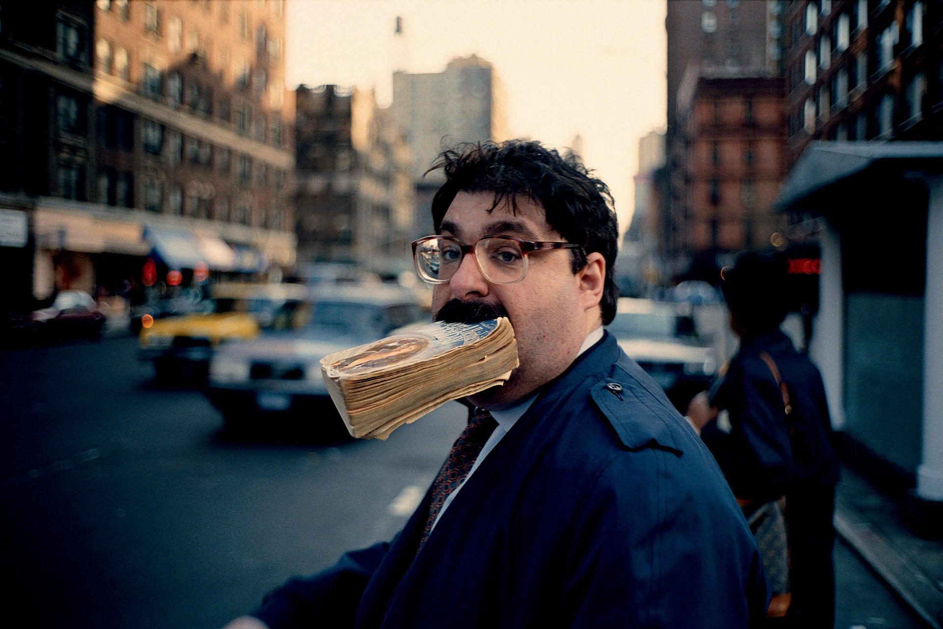 A man wearing glasses, with a moustache, tie and coat, walks across a New York street, holding a worn book in his mouth. In the background are passers-by, buildings, cars and a New York taxi.