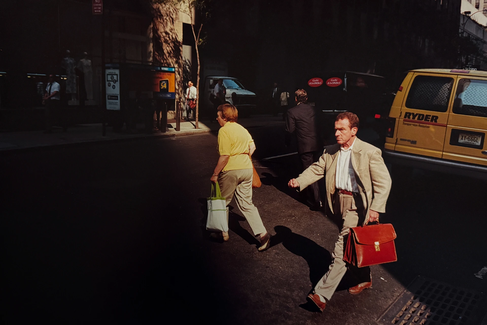 Uomo con valigetta portadocumenti rossa e donna con borsa della spesa che camminano su una strada di New York attraversando un raggio di sole.
