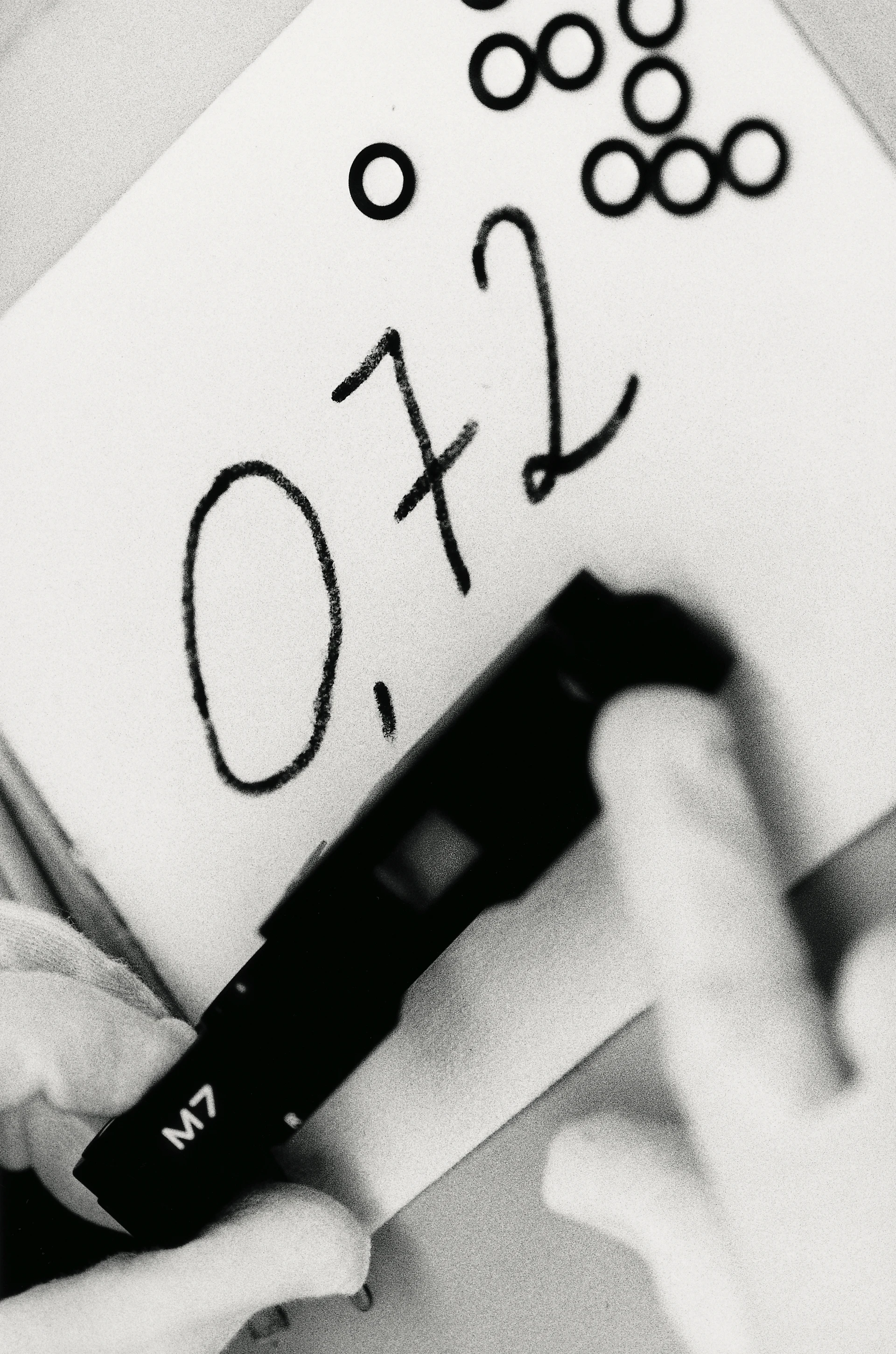 Primo piano in bianco e nero del coperchio superiore di una Leica M7 tenuto da mani con guanti davanti a un foglio di carta su cui è scritto in grande il numero 0,72.