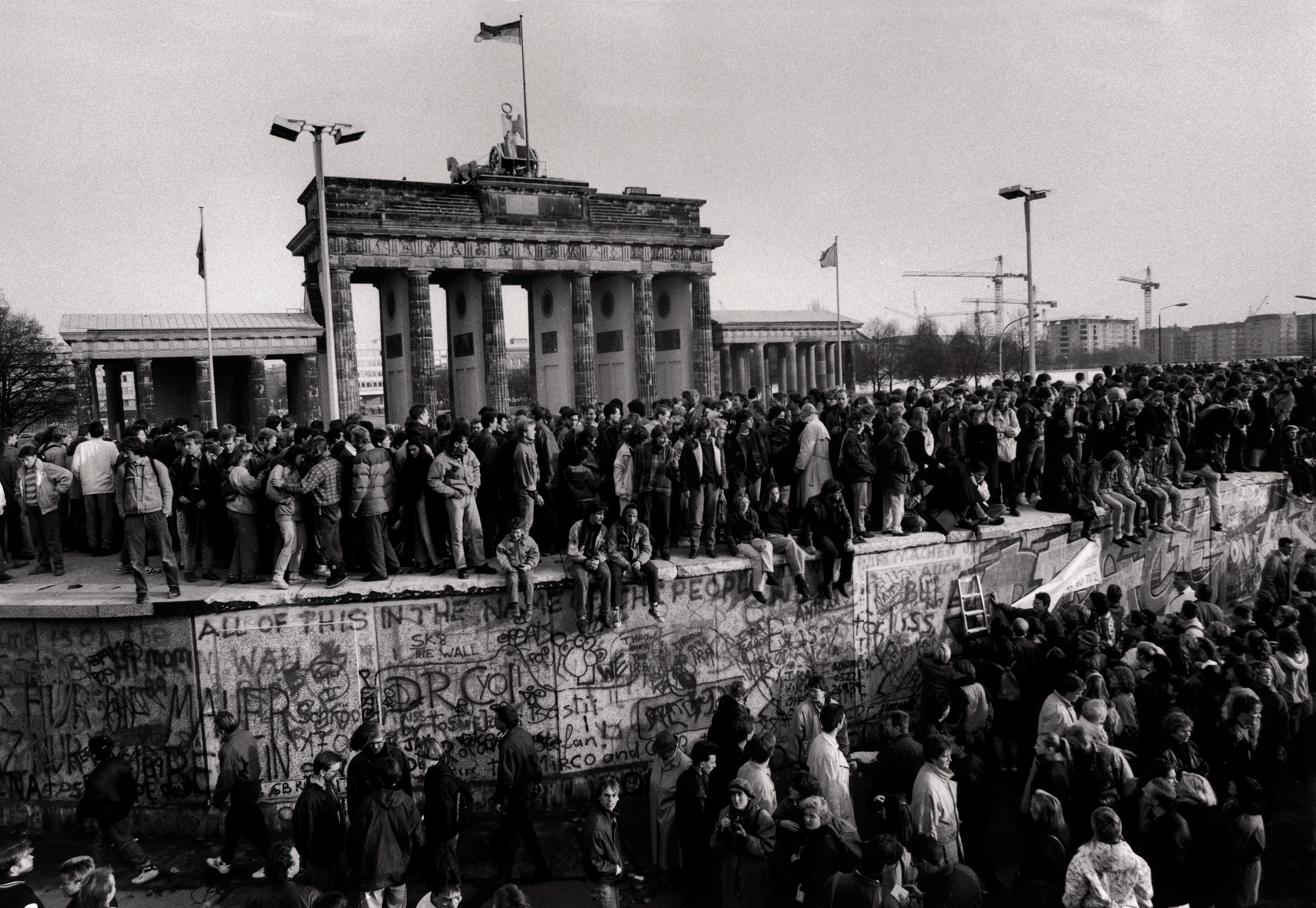 Foto in bianco e nero di una grande folla di persone radunate davanti e sopra il Muro di Berlino, sullo sfondo la Porta di Brandeburgo.