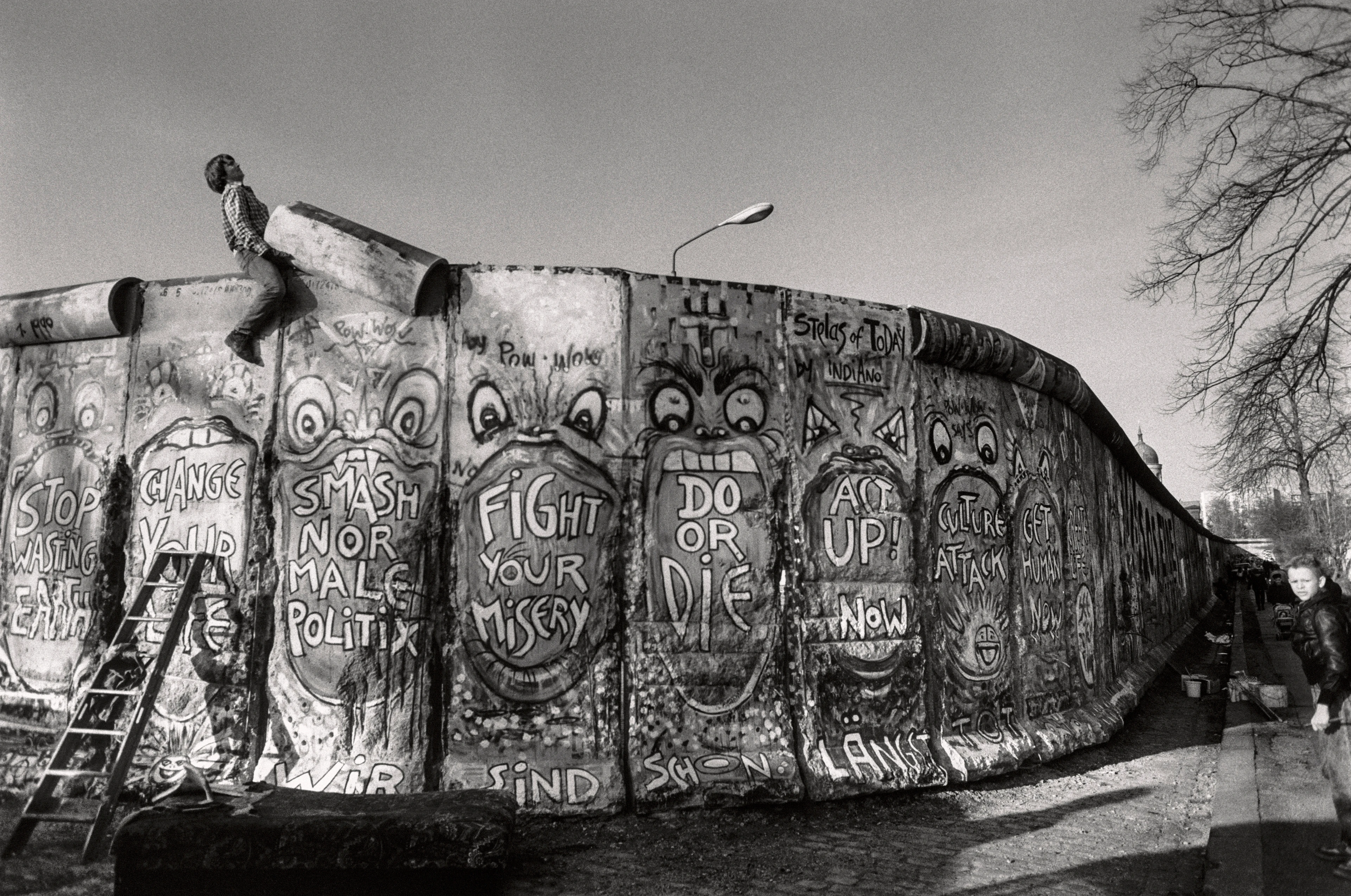 Foto in bianco e nero di una parte del Muro di Berlino, su cui sono dipinti numerosi messaggi, un uomo sta rimuovendo dall’alto la barriera in cemento.