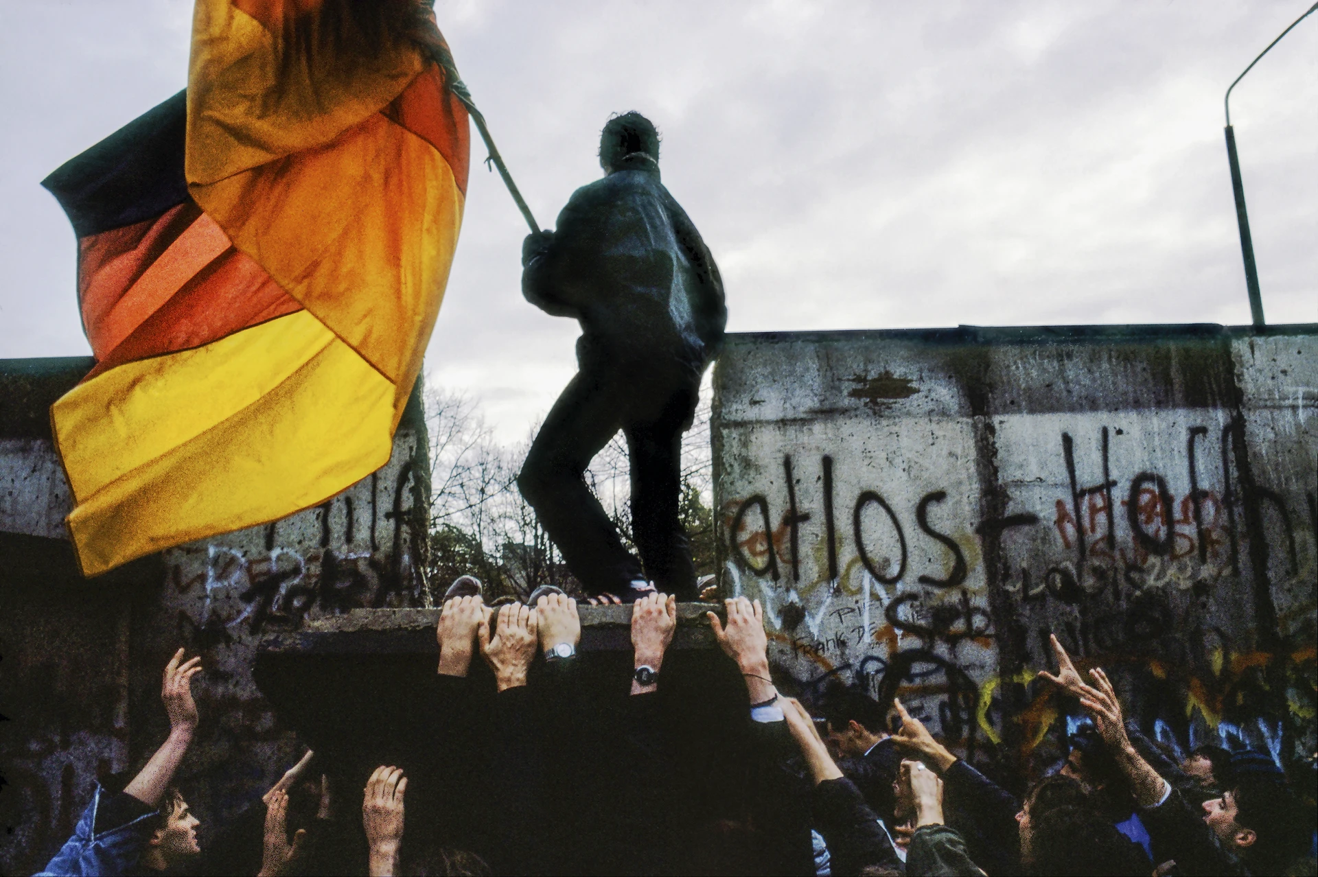 Photographed from within the crowd, people are seen destroying a section of the Berlin Wall. A man stands atop the demolished portion, holding a large German flag, while numerous hands support the broken structure beneath him.