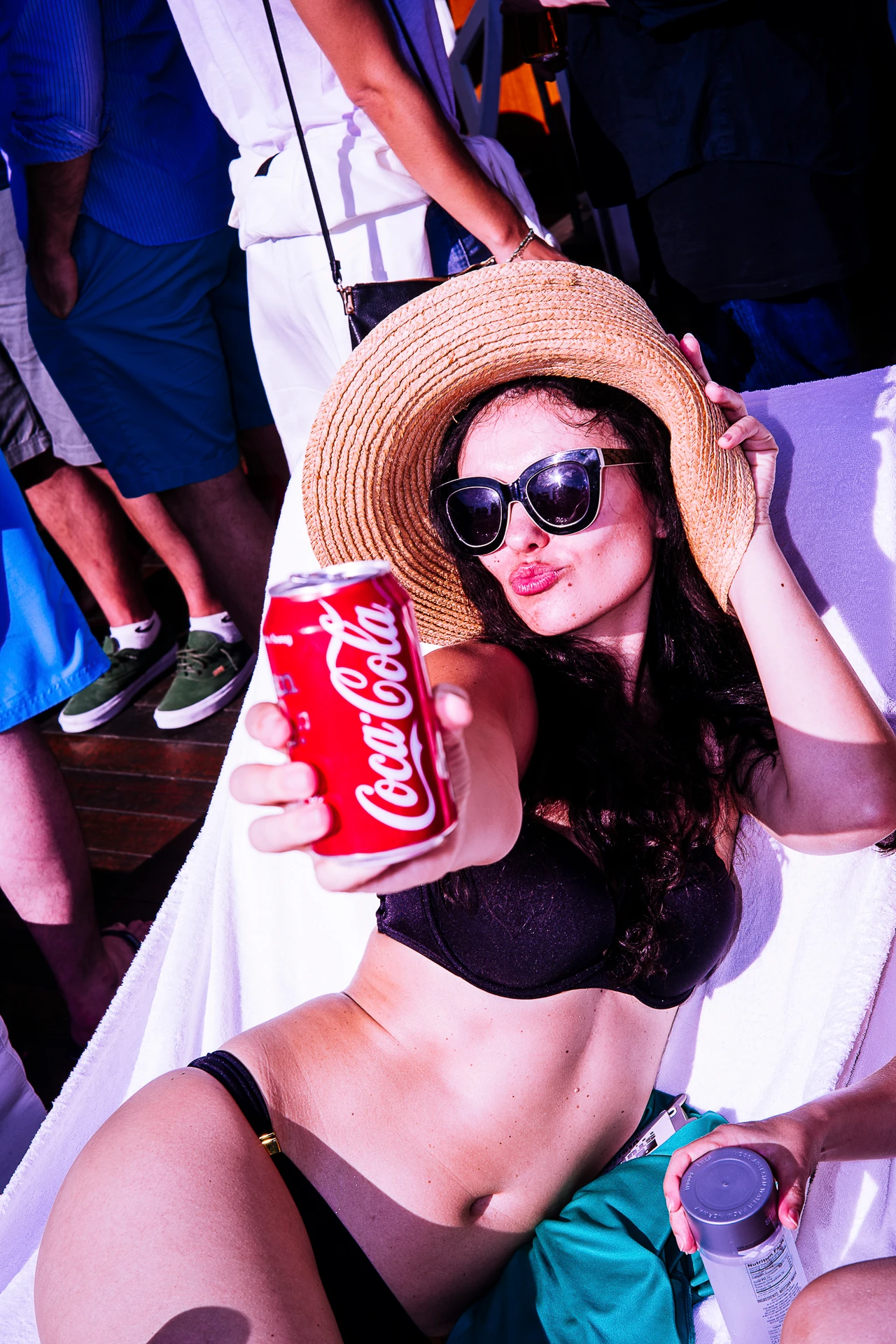 A young woman wearing a sun hat and sunglasses holds a red drinks can and looks into the camera, making a kissy face for the camera while other people stand close around her.