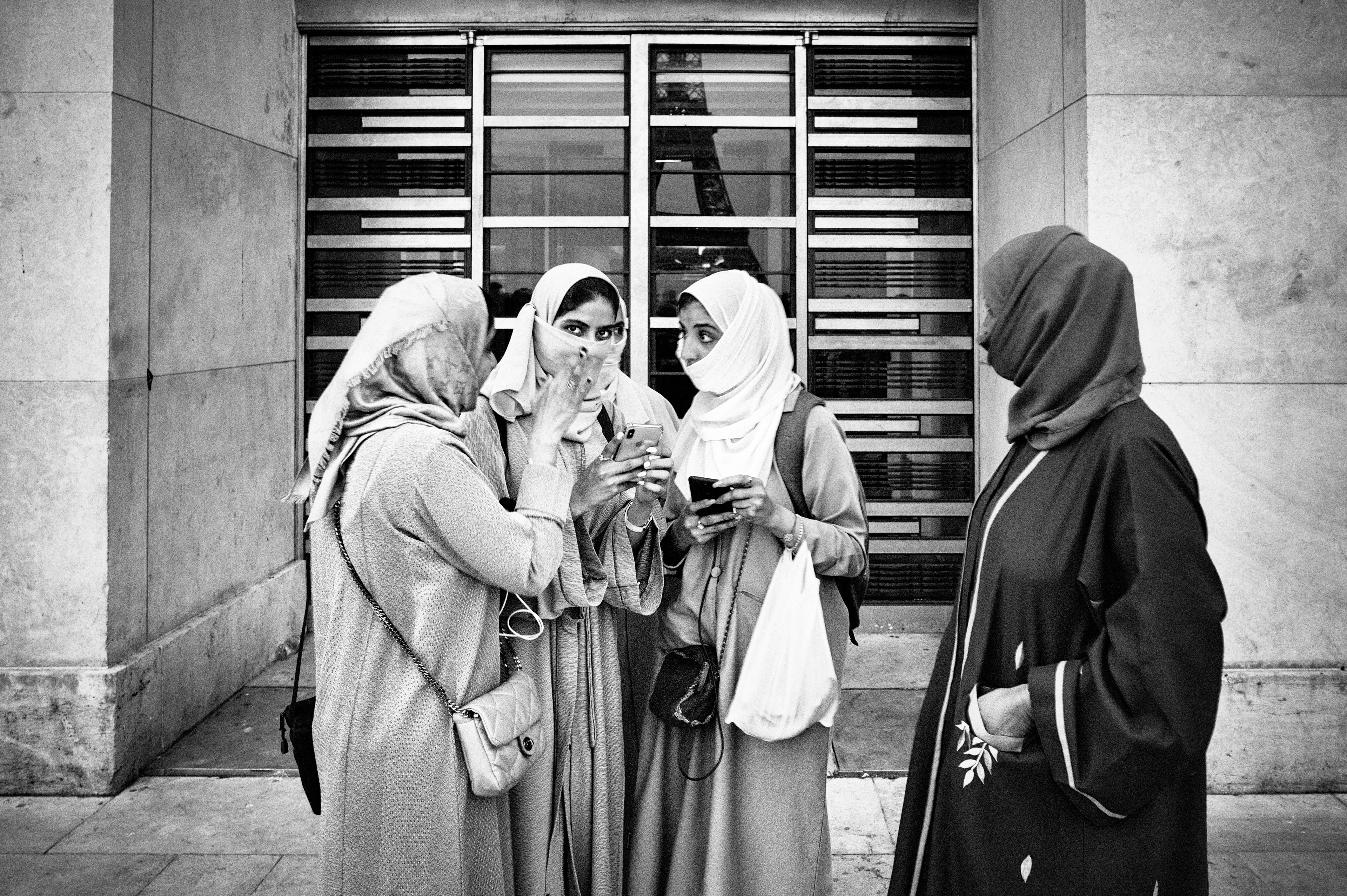Black-and-white image: Four women wearing headscarves stand close together in front of a building, holding smartphones. One of them is looking directly into the camera while another person in the foreground looks curiously at the setting.