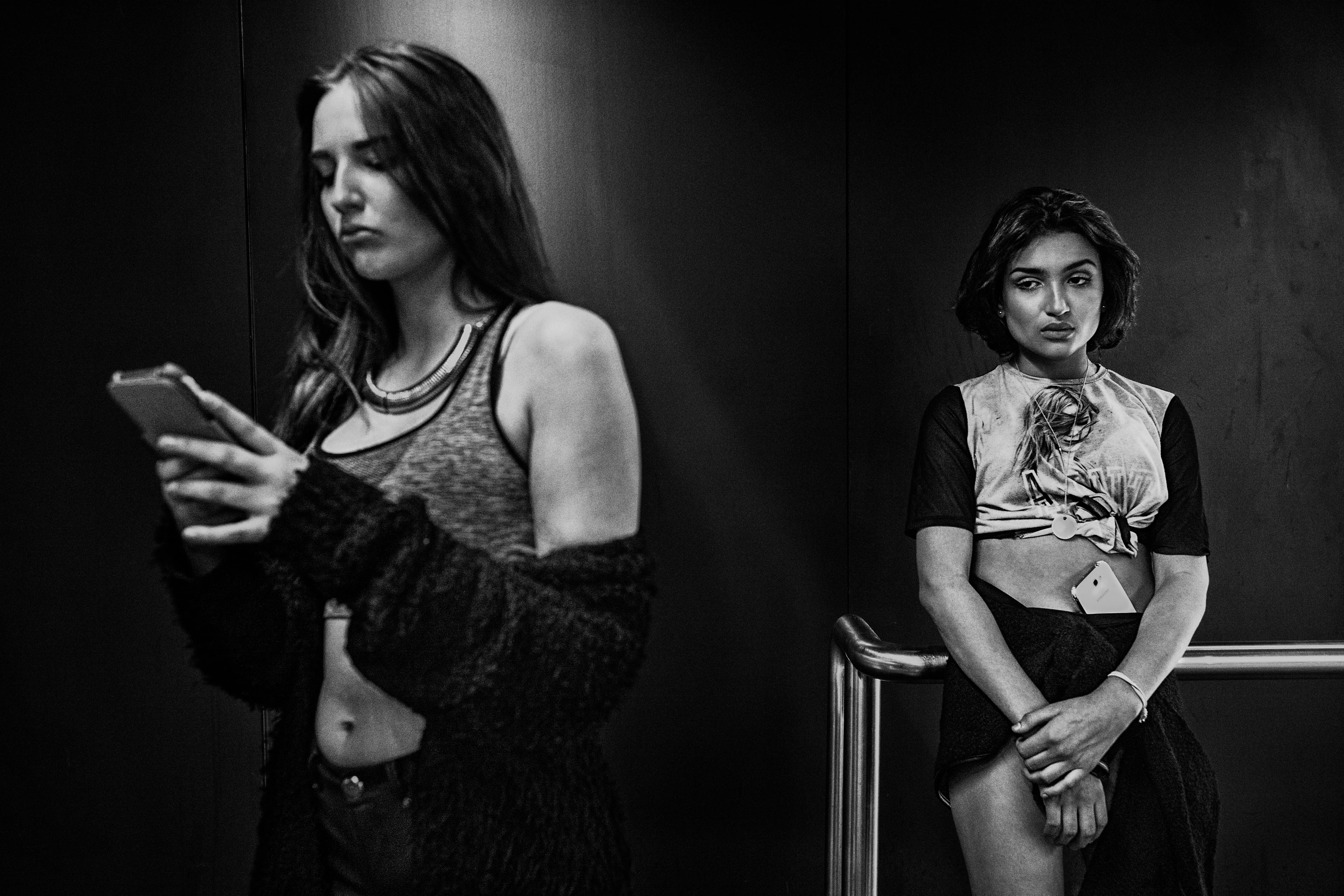 Black-and-white image: Two women stand in front of a black wall. The young woman on the left looks at her mobile phone, while the young woman on the right leans against a railing, staring into space with her smartphone tucked into her skirt at navel height.
