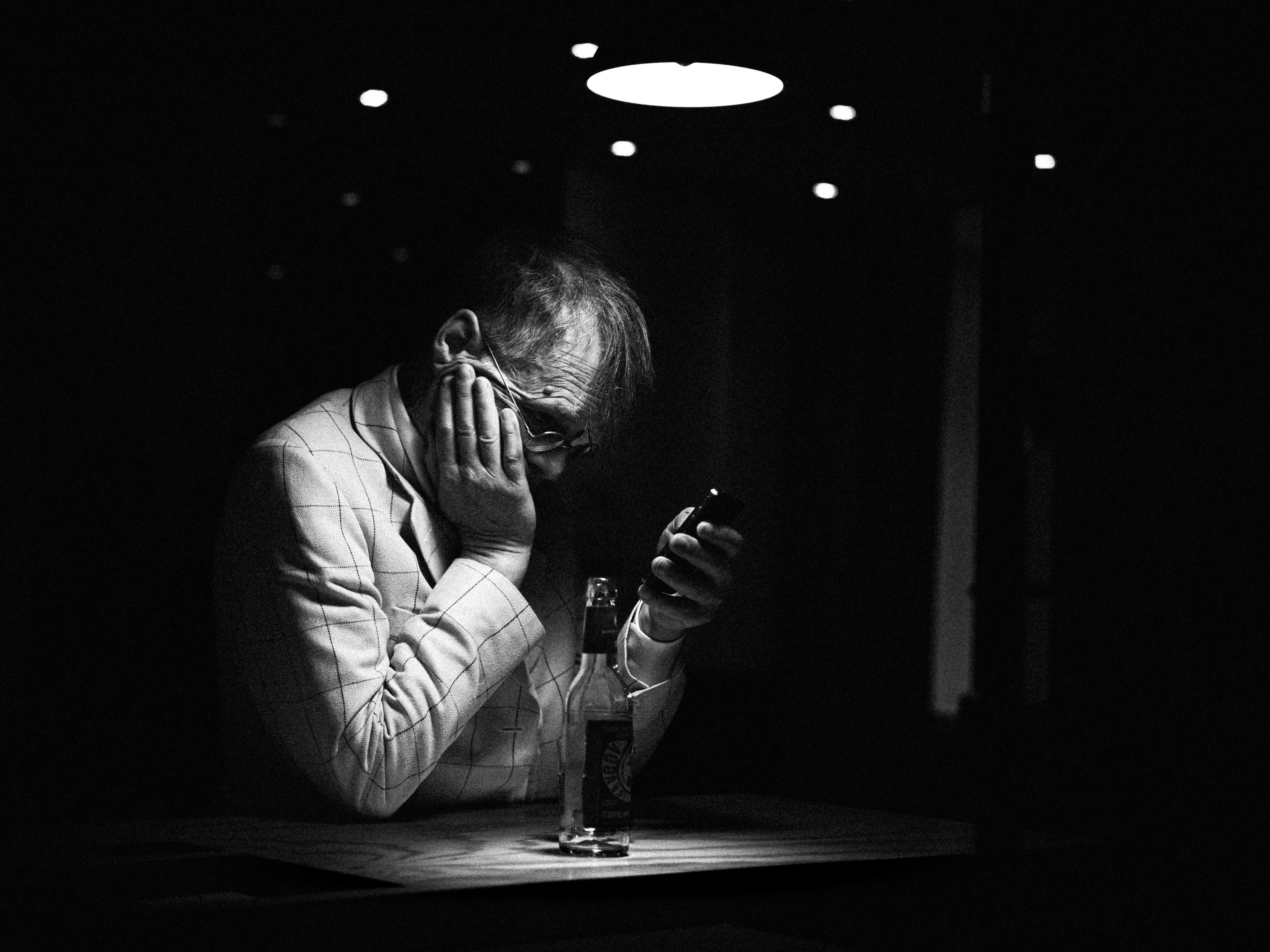 Black-and-white image: A man sits in a dark room in a cone of light, resting his head in his hand as he looks fixedly at his smartphone. An empty beer bottle stands on the table in front of him.