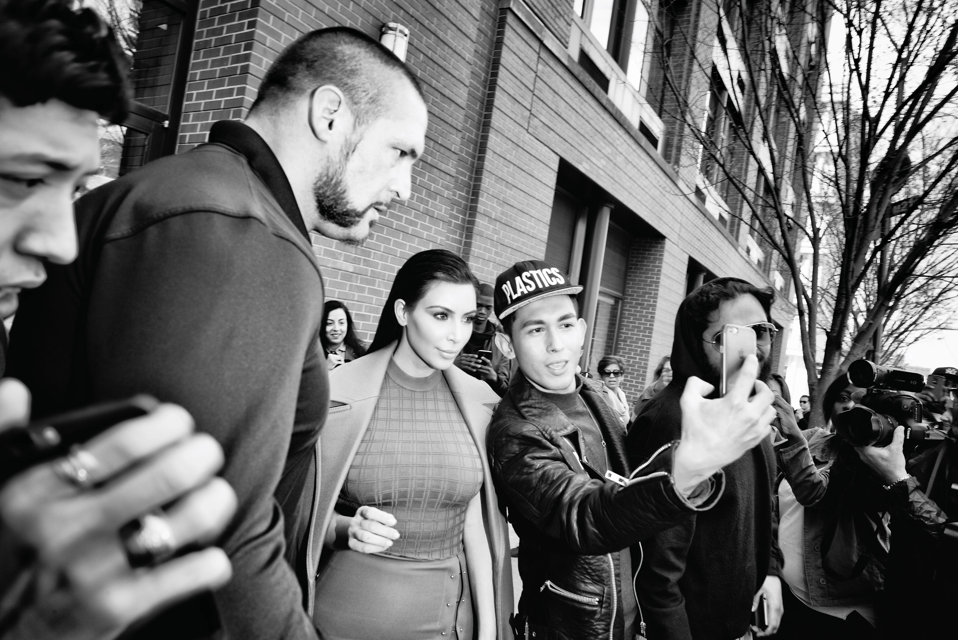 Black-and-white image: Kim Kardashian is walking down the street, accompanied by a bodyguard and surrounded by onlookers and paparazzi. A man wearing a baseball cap takes a selfie with her; she looks confidently into his camera.