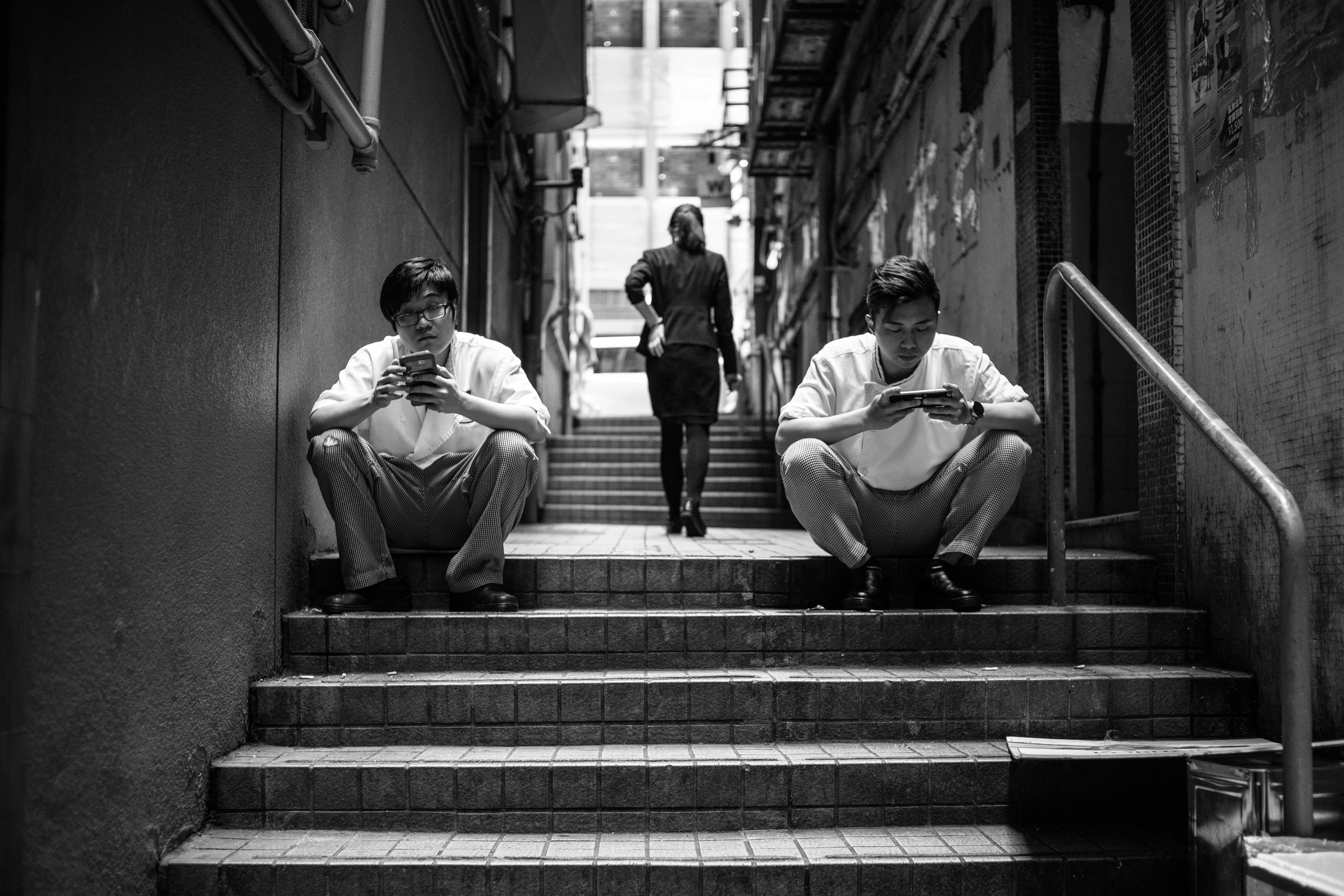 Black-and-white image: Two young men sit on a staircase, each looking at their smartphone, while a woman walks up the stairs in the background.