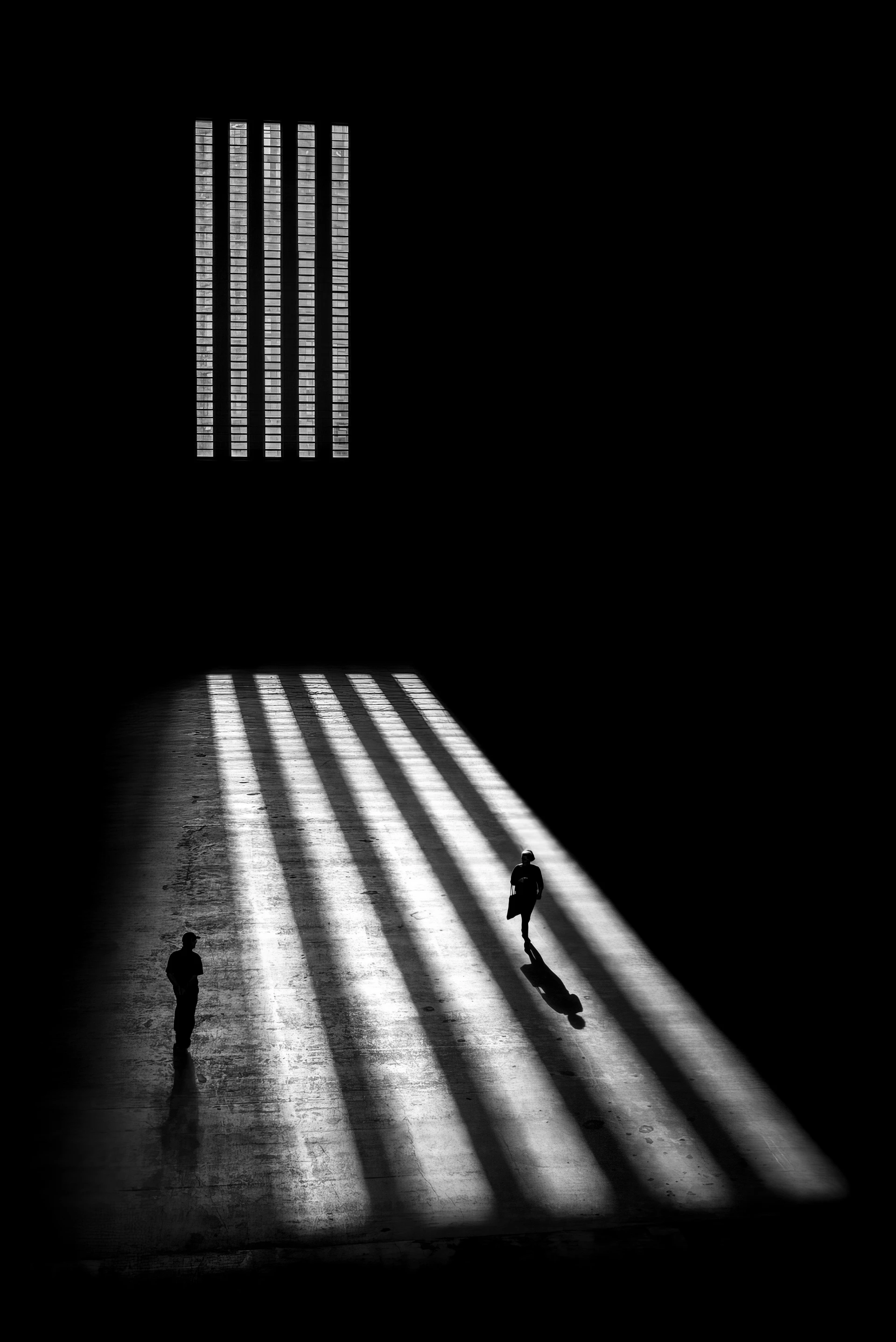 有氛围感的黑白照片:明亮光线形成的斜射条纹贯穿巨大的黑暗大厅,一个人正穿行其间,而另一个人则立于光线之外。