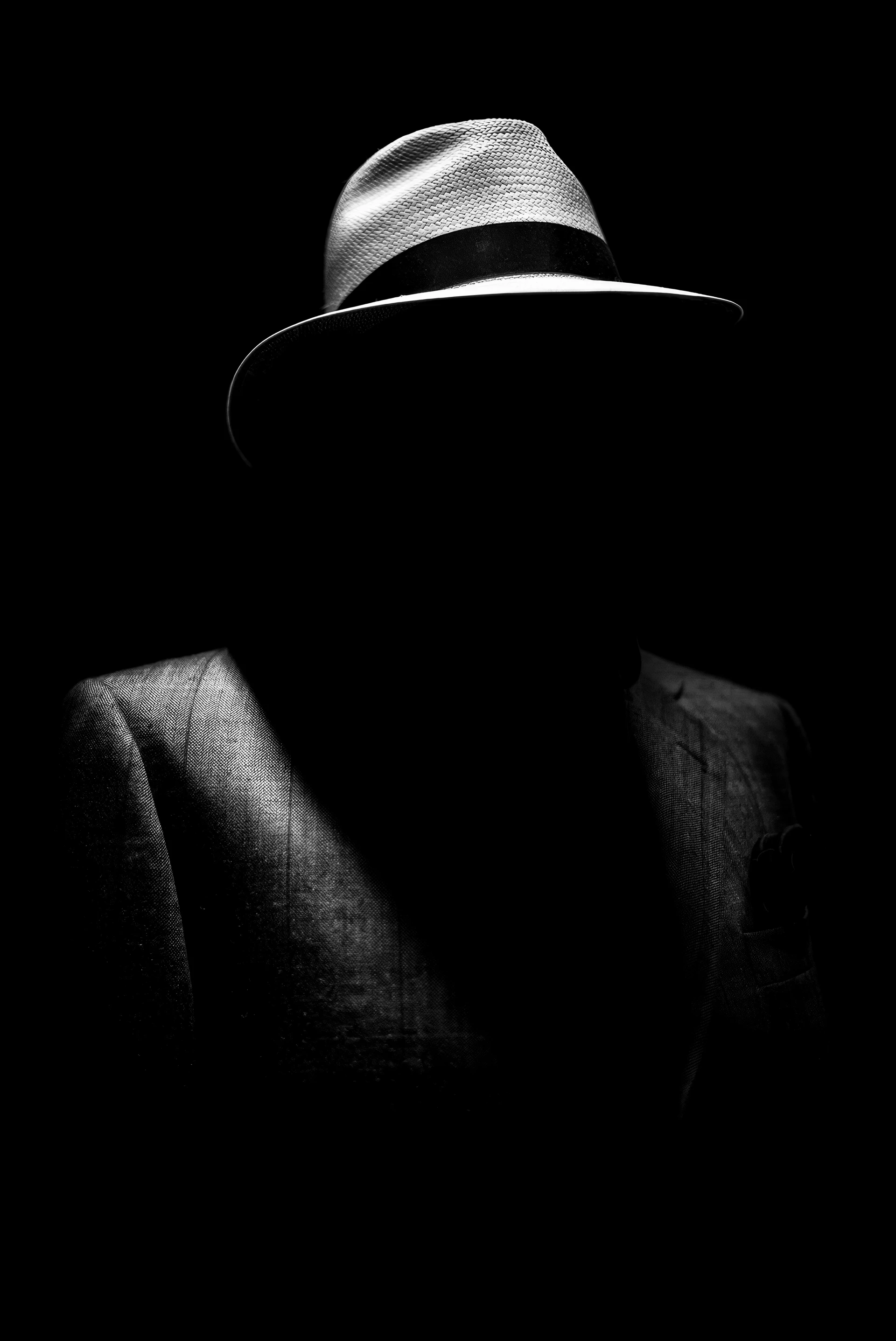 Dark black-and-white image: A person wearing a hat stands in the dark. Their face is completely in shadow, so only their hat and shoulders of their coat are visible.