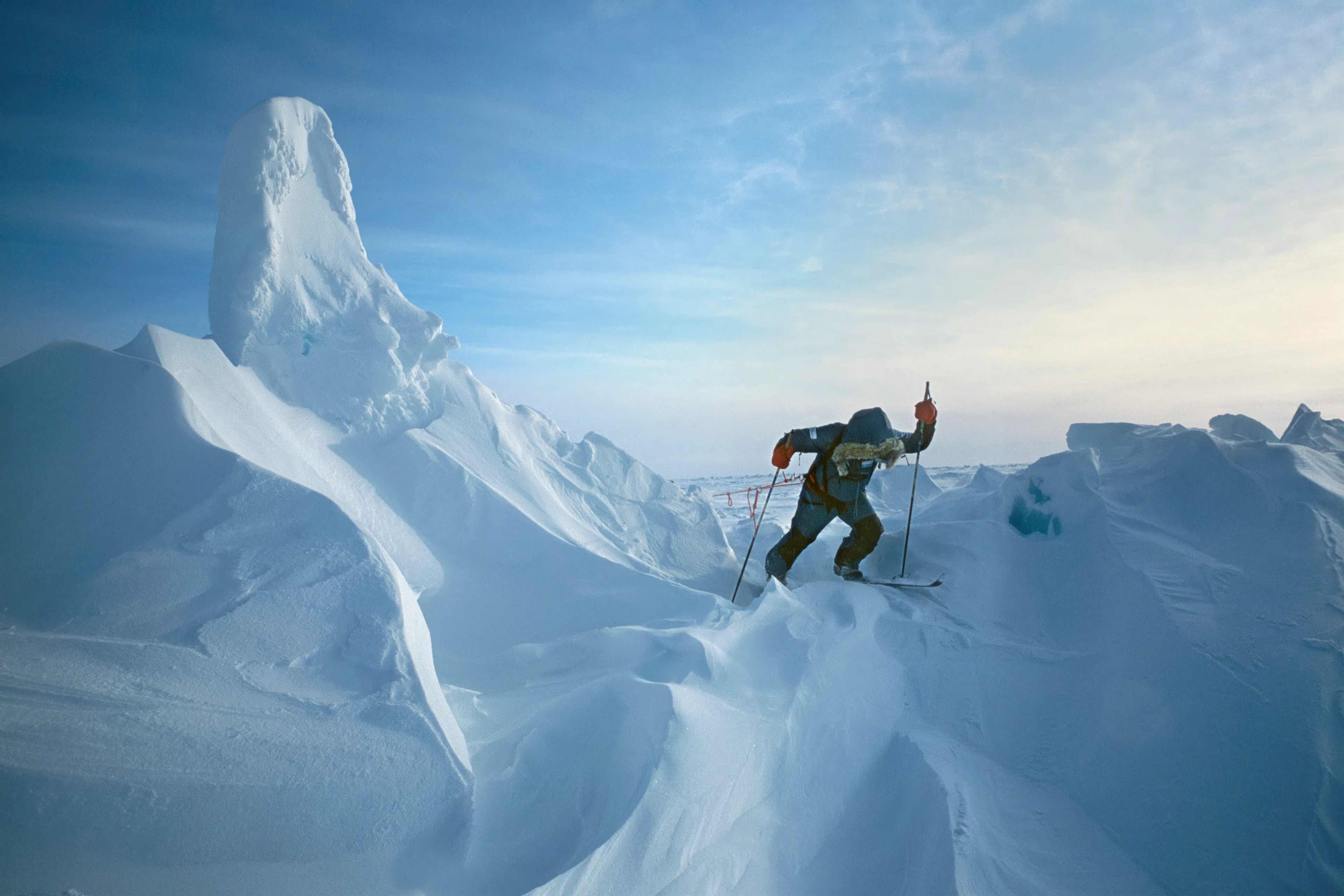 A lone polar explorer pulls a fully loaded sledge on skis across rugged snowfields characterised by high ice formations, under a clear, cool Arctic sky.