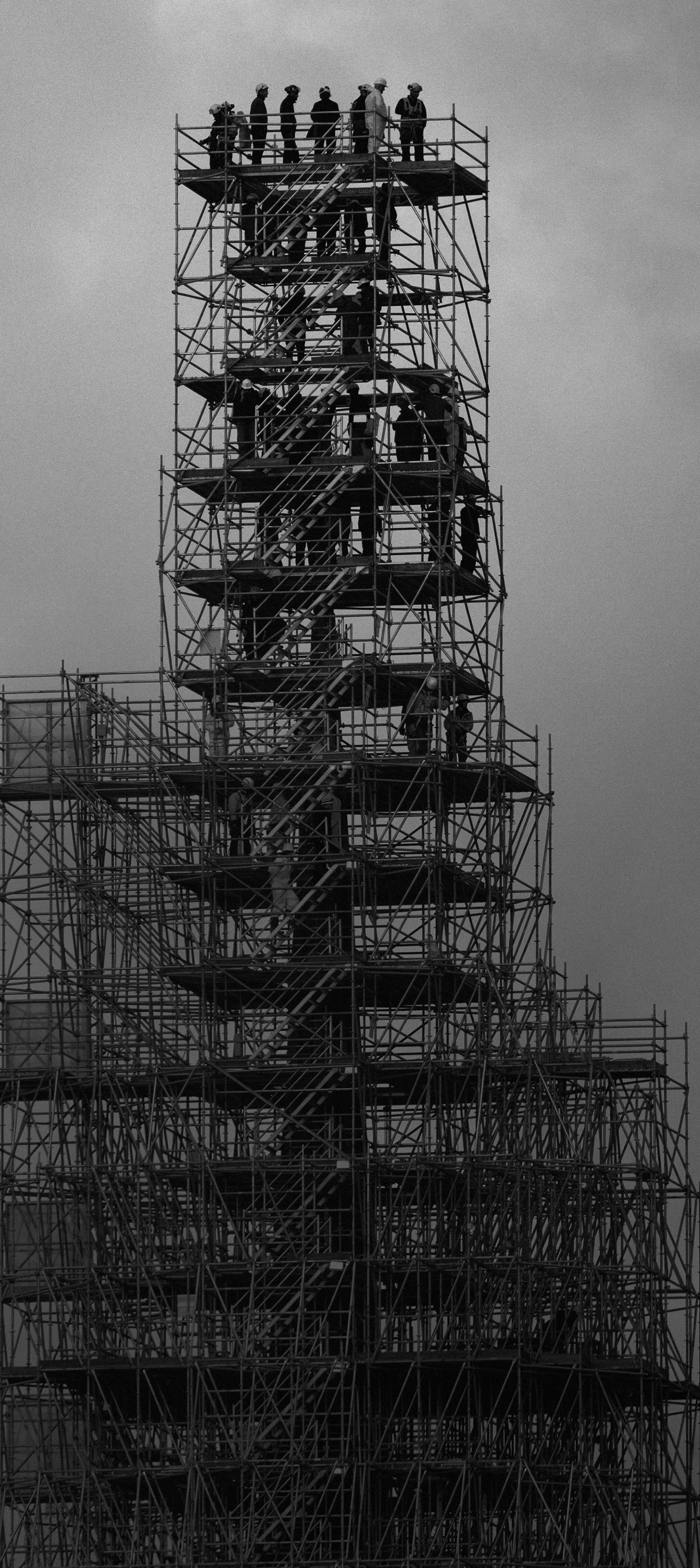 Schwarz-Weiß-Bild: Ein hoher Turm aus Baugerüsten mit zahlreichen Arbeitenden, die sich auf verschiedenen Ebenen bis zur Spitze hin bewegen; der Hintergrund besteht aus bewölktem Himmel.