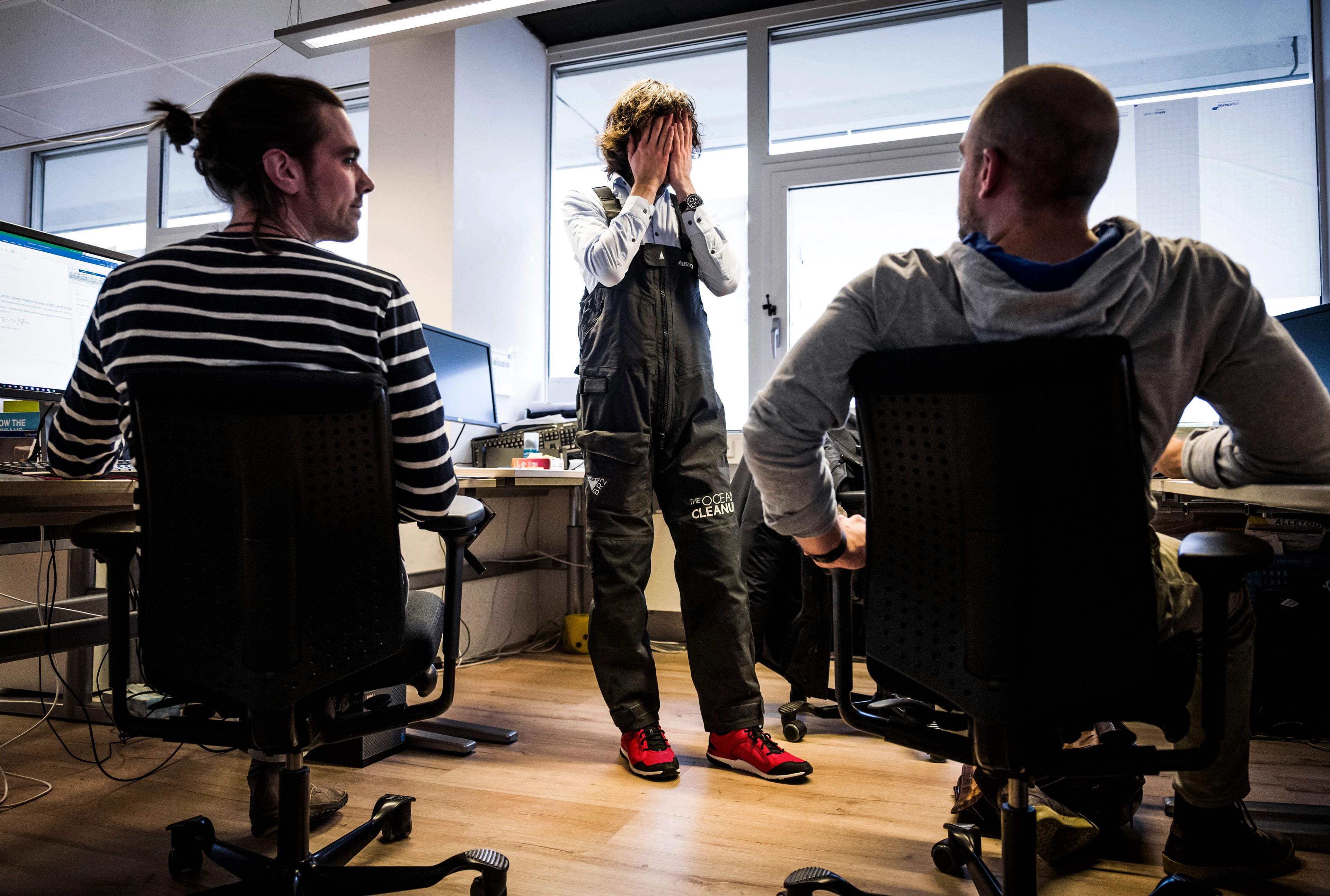Boyan Slat, wearing black overalls bearing the name of his company, The Ocean Cleanup, stands in the office in front of two colleagues sitting in chairs, covering his face with his hands.