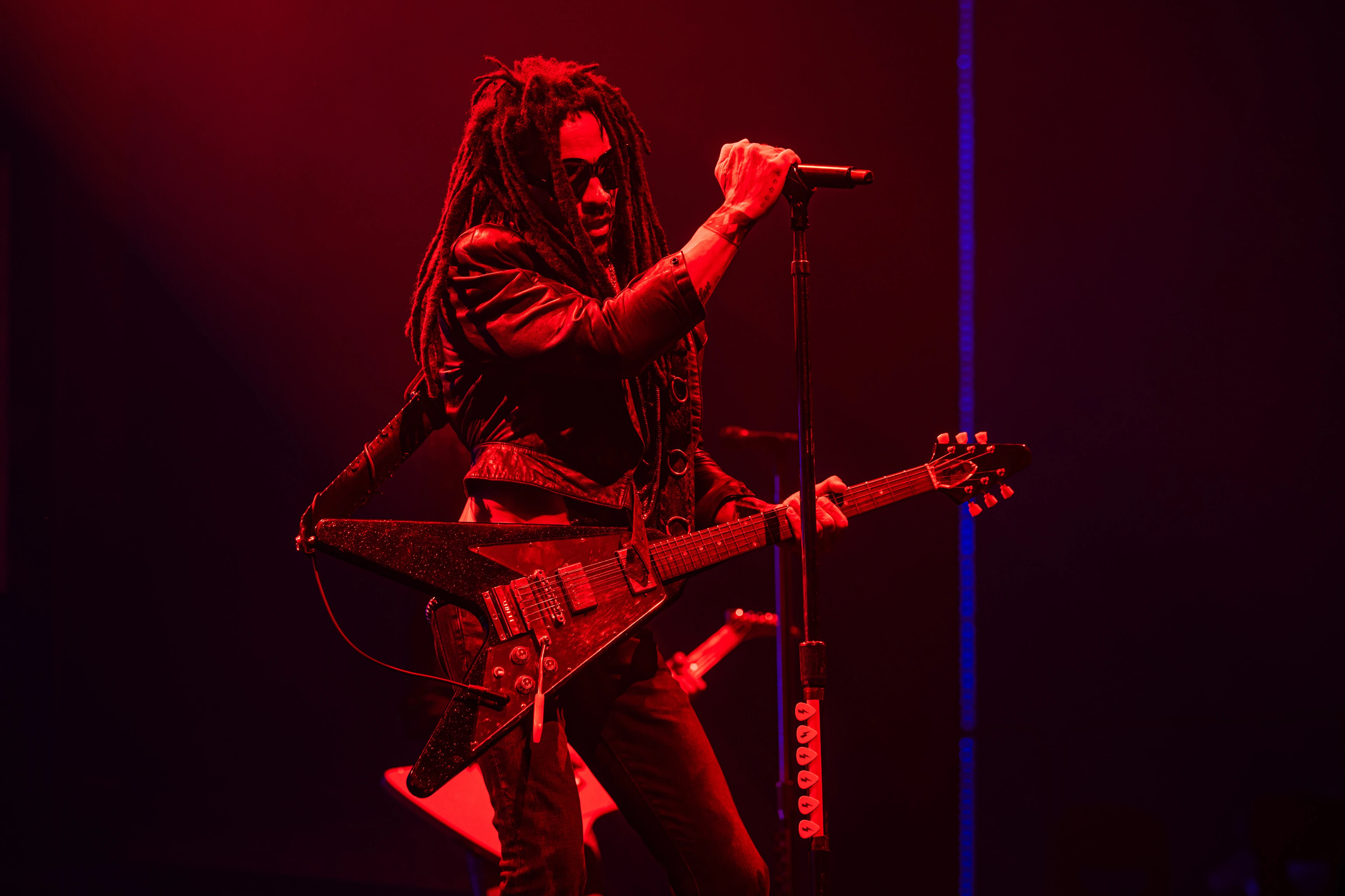 A stage bathed completely in red light with two delicate neon blue LED strips in the background. Lenny Kravitz stands with a cool stance in the red spotlight, one hand on the microphone, sunglasses on his face and an electric guitar slung over his shoulder.