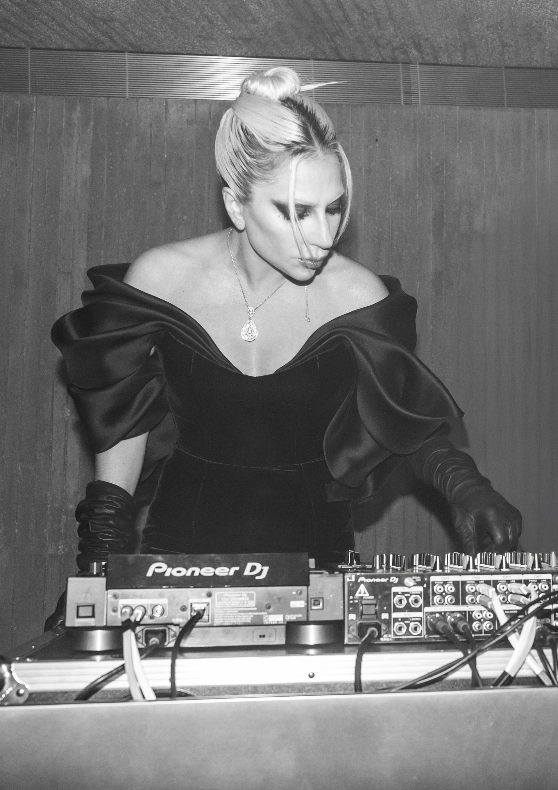 Schwarz-Weiß-Bild: Die konzentrierte Lady Gaga steht beim Auflegen in schwarzem Abendkleid hinter einem DJ Mischpult.