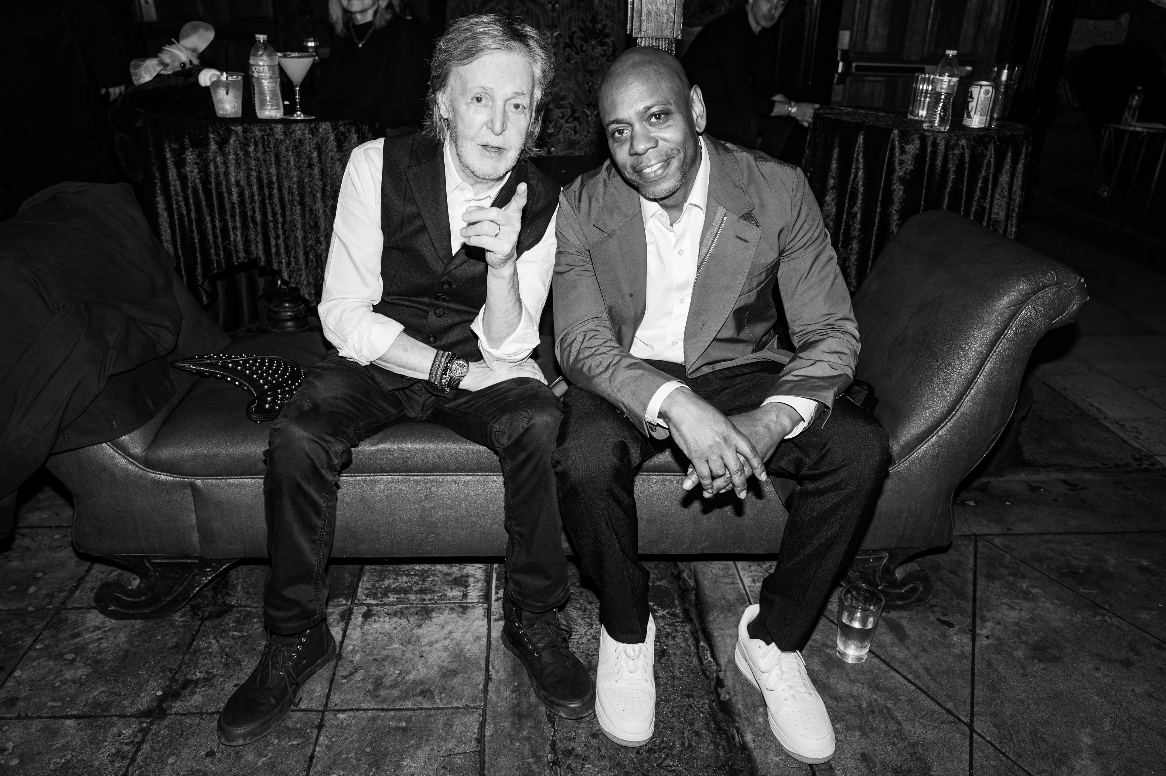 Schwarz-Weiß-Bild: Paul McCartney und Dave Chappelle sitzen Knie an Knie auf einer Chaiselongue und posieren lächelnd für die Kamera.