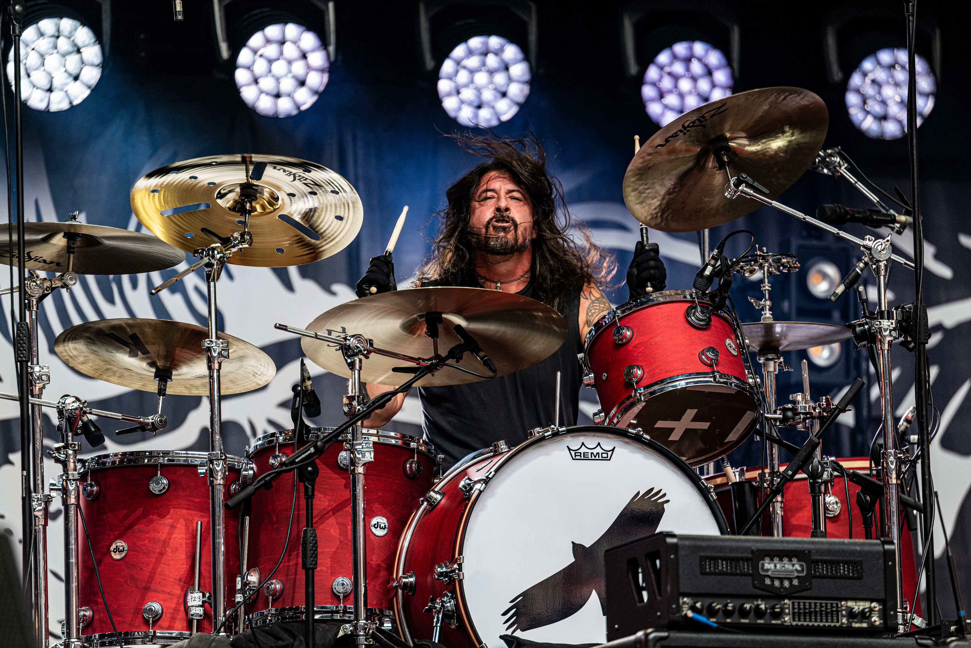 Dave Grohl in Aktion, er sitzt hinter einem roten Schlagzeug auf der Bühne, die Stöcke und Haare fliegen während er wild entschlossen Richtung Kamera blickt.
