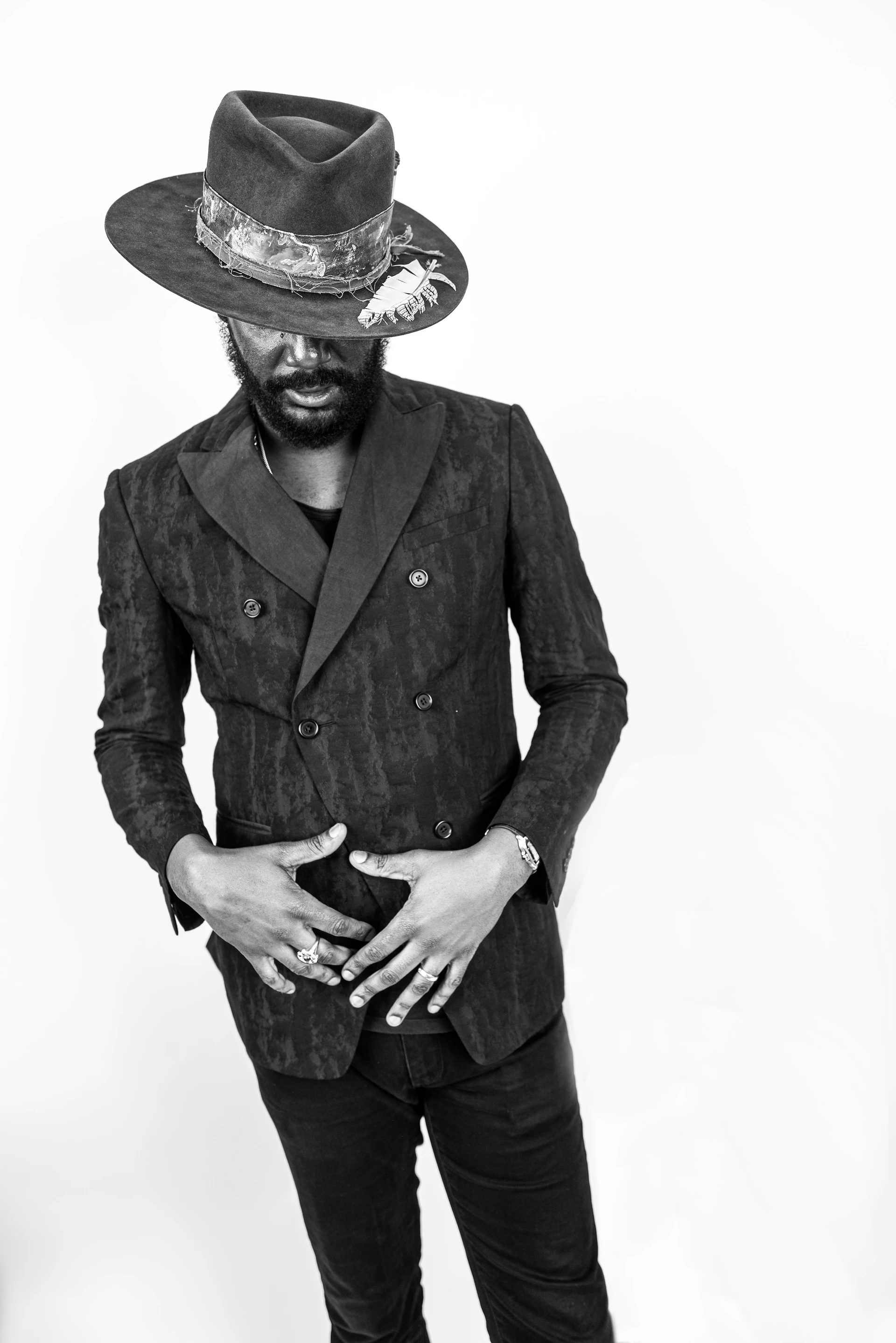 Schwarz-Weiß-Porträt: Vor weißem Hintergrund steht Gary Clark Jr. im schwarzen Anzug und Hut, er blickt nach unten und hält beide Hände ausgebreitet vor sein Sakko.