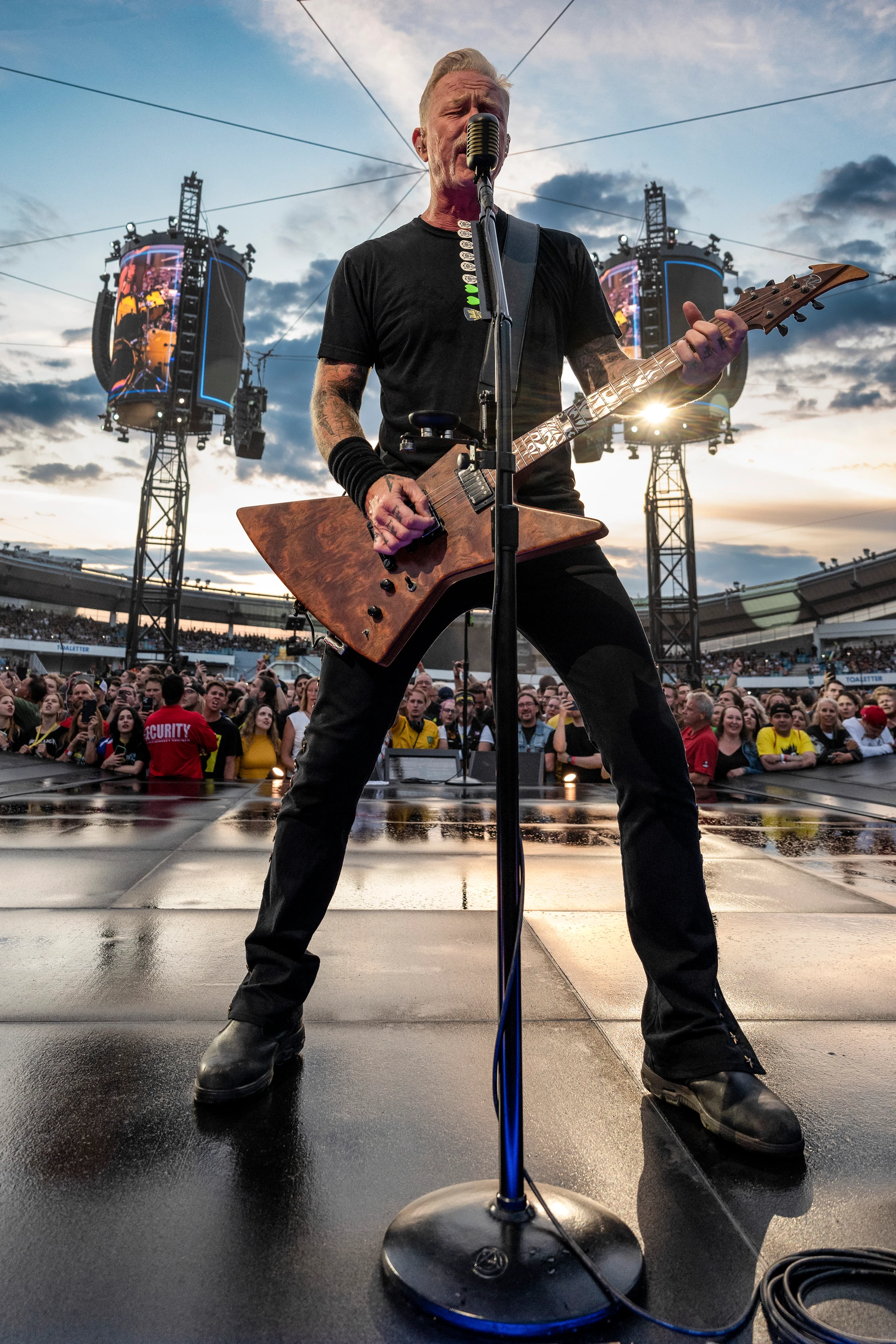 Schwarz-Weiß-Porträt: James Hetfield von Kniehöhe aus nach oben gegen den Himmel fotografiert, auf einer Bühne mit E-Gitarre, er singt in ein Mikrofon, während im Hintergrund ein großes Publikum und Stadionbeleuchtung zu sehen sind.