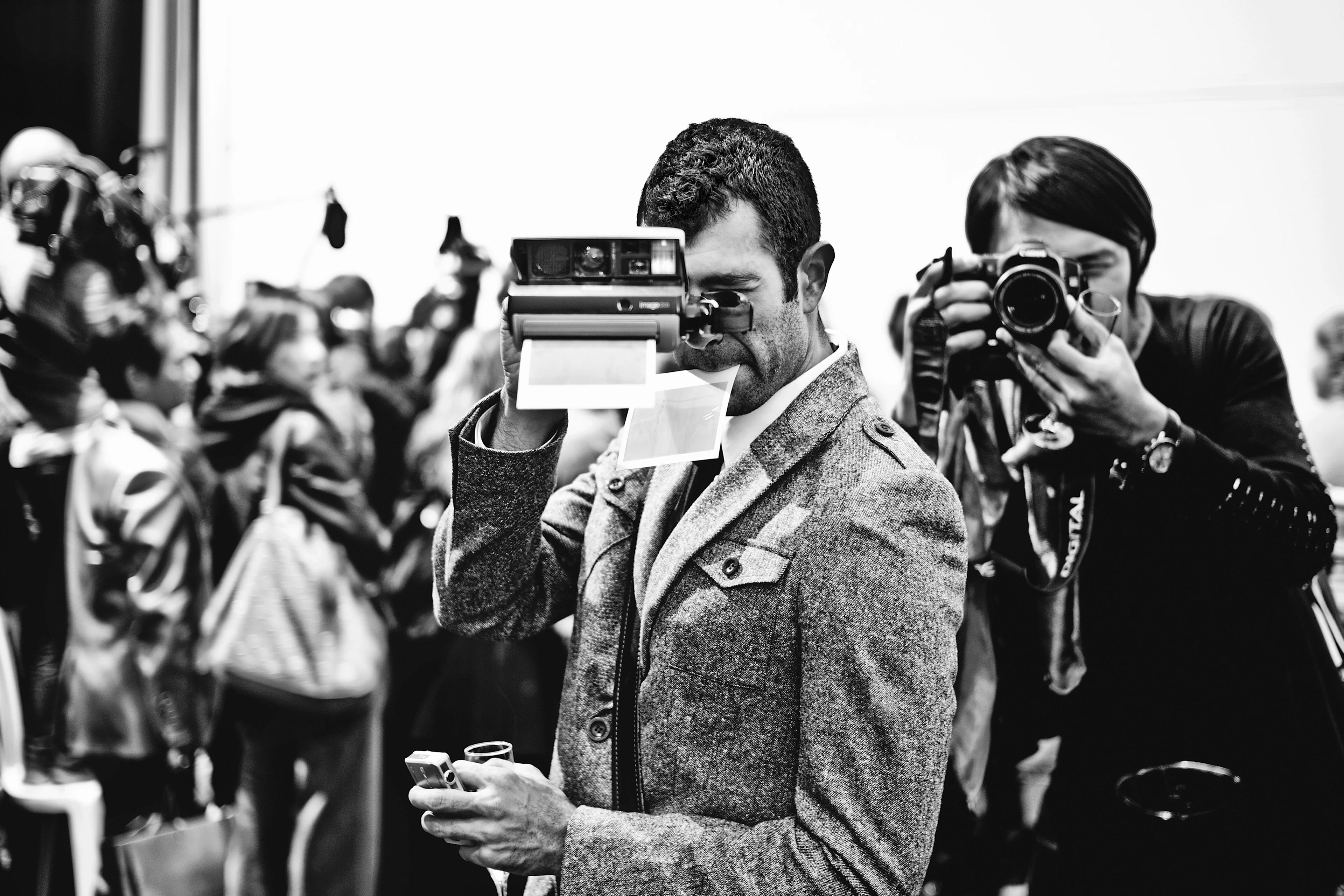Black-and-white image: Two photographers stand in the foreground, facing the camera. One is taking pictures with an instant camera and the other with a compact camera. A cluster of blurred journalists can be seen in the background.